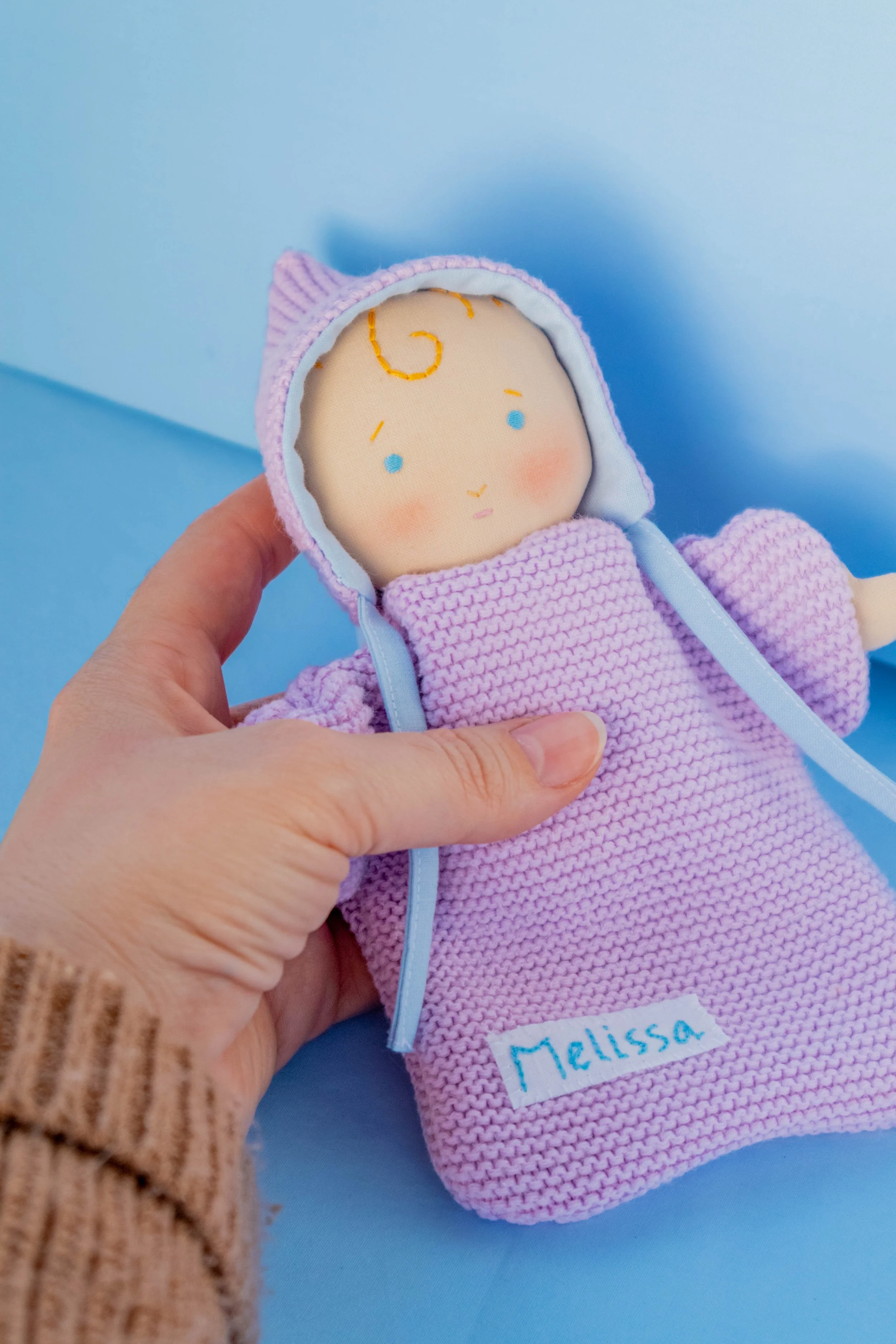 A handmade doll with a embroidered face, wearing a purple hooded outfit, held by a person's hand against a blue background. The doll has a label with the name "Mellissa."