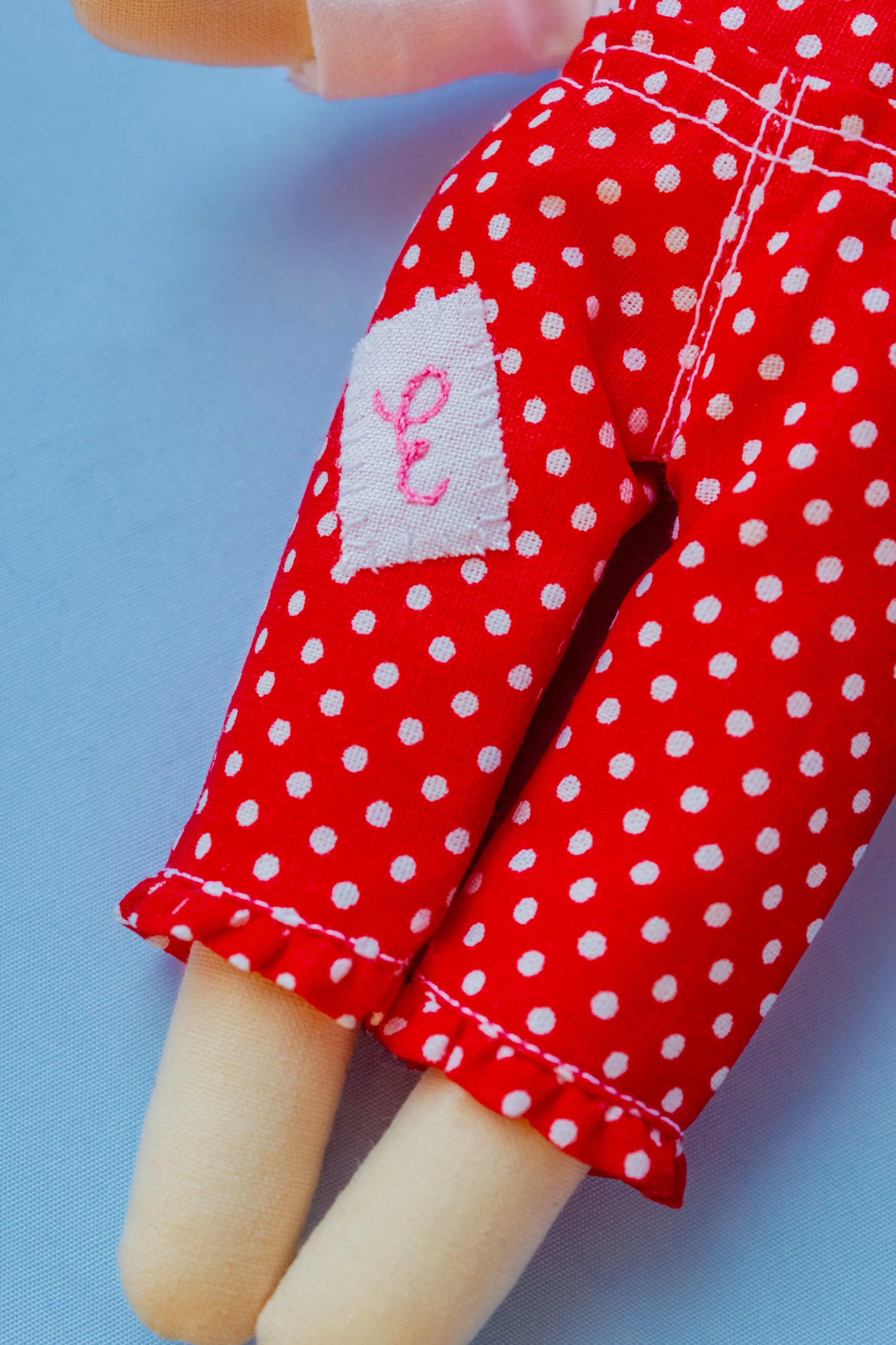 Close-up of a doll's red polka dot dress with a white patch and pink embroidery, beige-colored legs, and a light blue background.