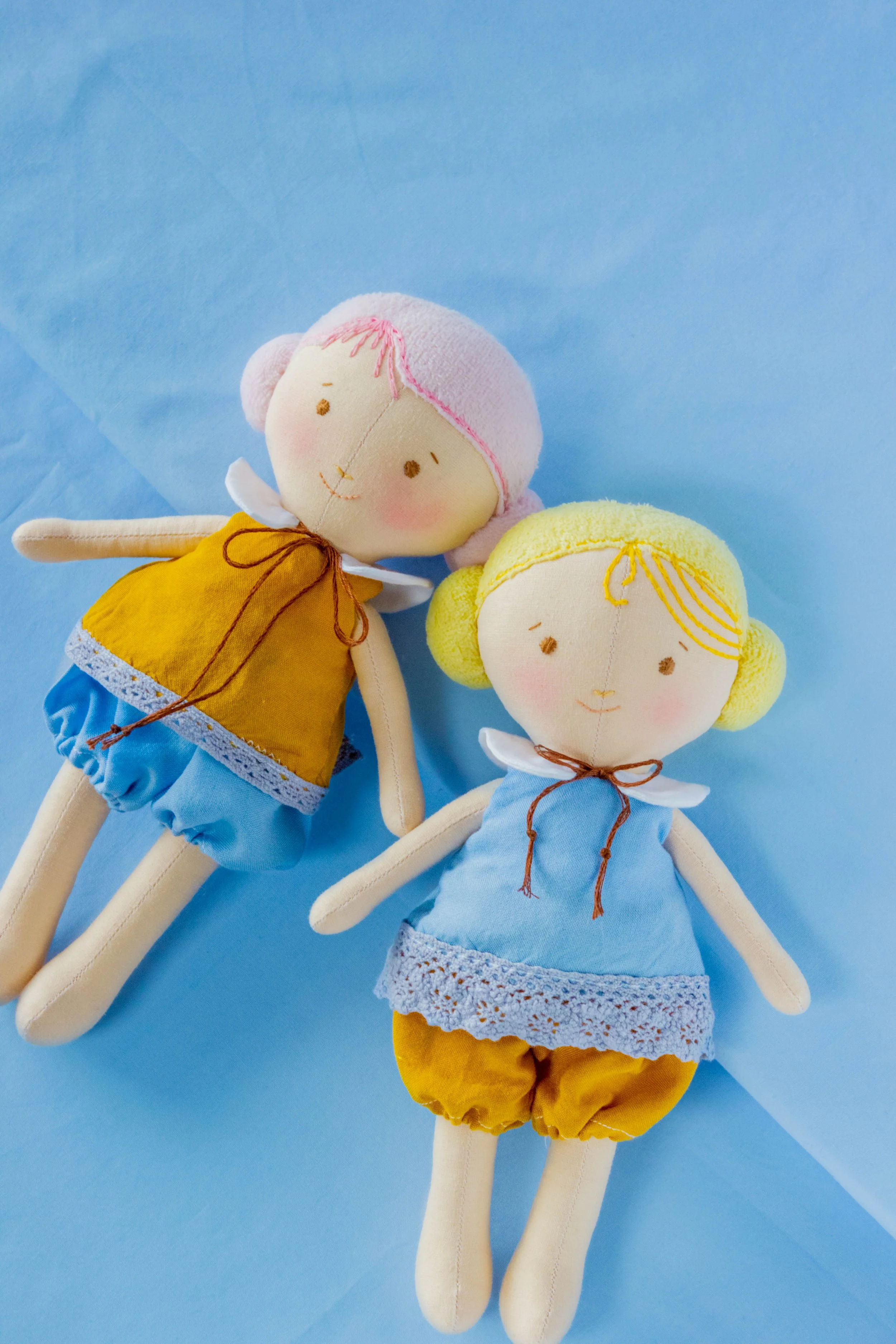 Two handmade cloth dolls lying on a blue surface, one with pink hair and the other with yellow hair, both wearing yellow and blue outfits with lace details.