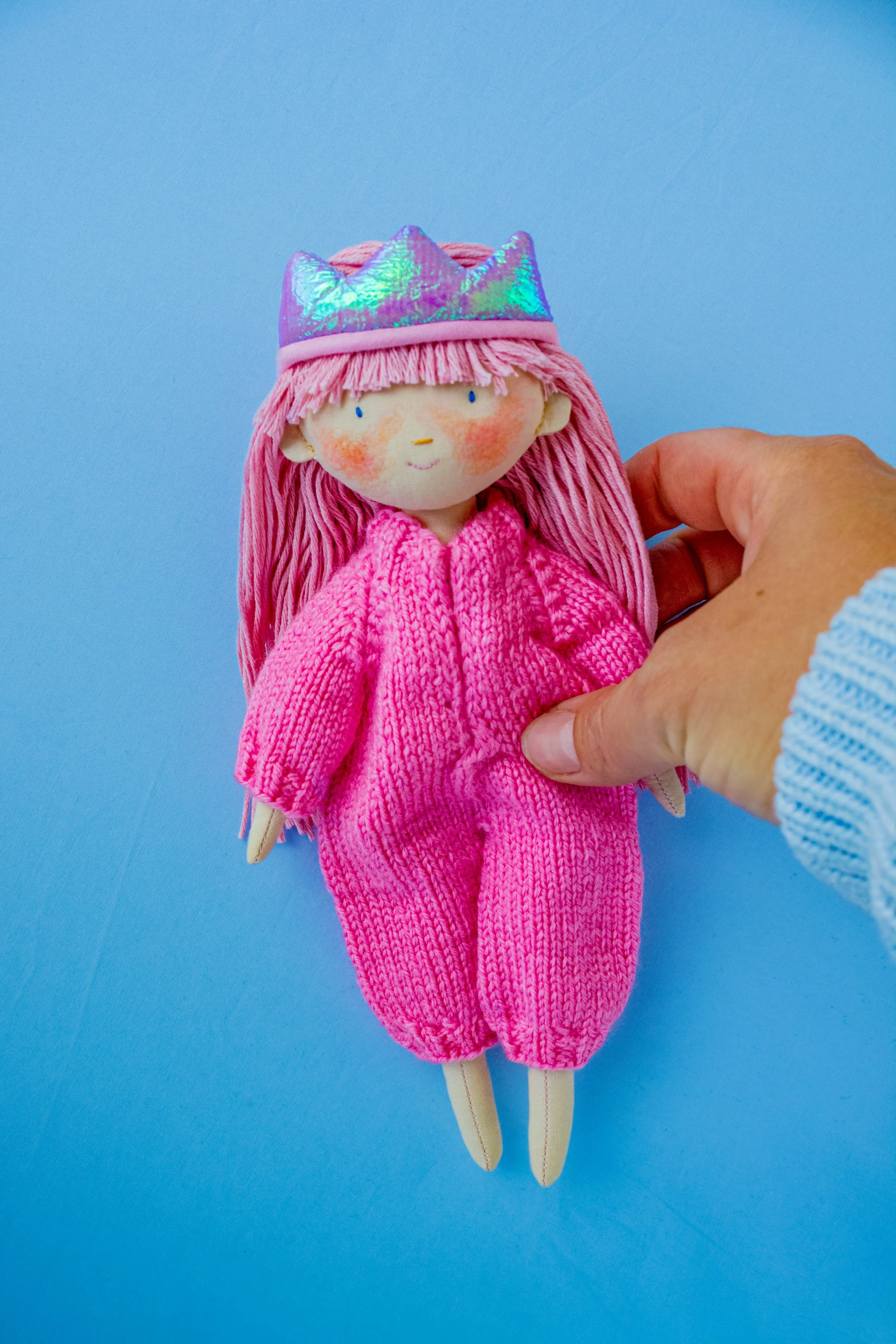 Hand holding a plush doll with pink hair, a pink knit outfit, and a shiny iridescent crown against a solid blue background.