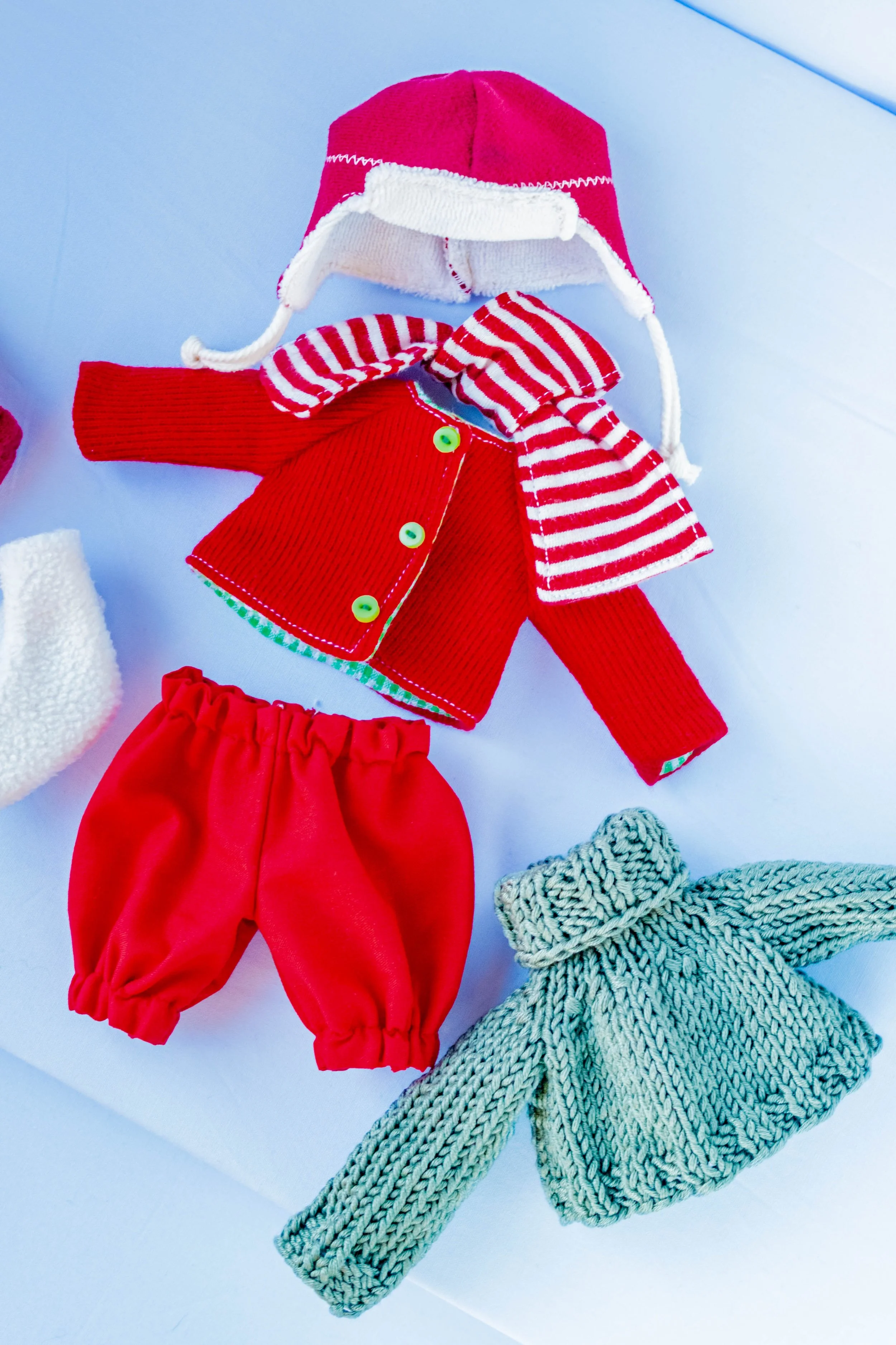 A collection of Christmas-themed clothing and accessories including a red and white Santa hat, a red jacket with green buttons, red pants, a red and white striped scarf, a white fuzzy hat, and a knitted gray scarf, all laid out on a light surface.