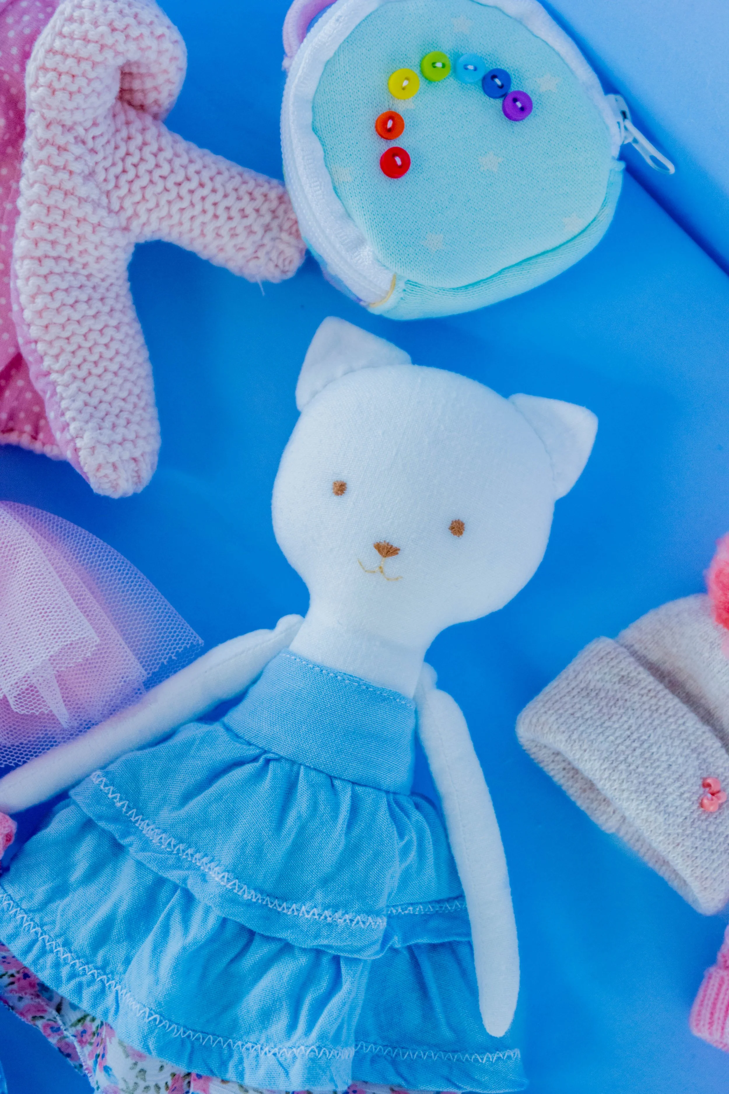 Close-up of a plush stuffed animal bear, dressed in a blue dress with ruffles and a white bunny toy, surrounded by pink knitted and fabric toys, against a blue background.