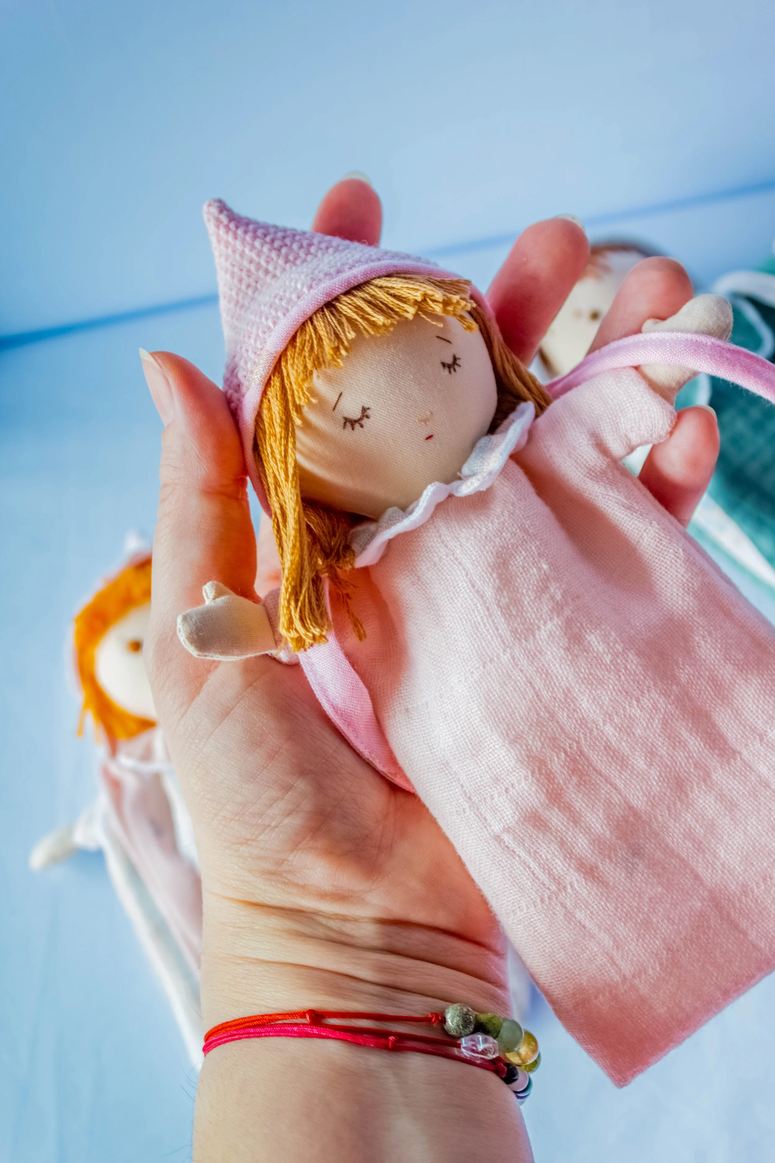 A person holding a handmade doll with orange hair, closed eyes, and a pink hat with a pointed top. The doll wears a pink dress with a ruffled collar. The person is wearing multiple colorful bracelets and a red string bracelet on their wrist.
