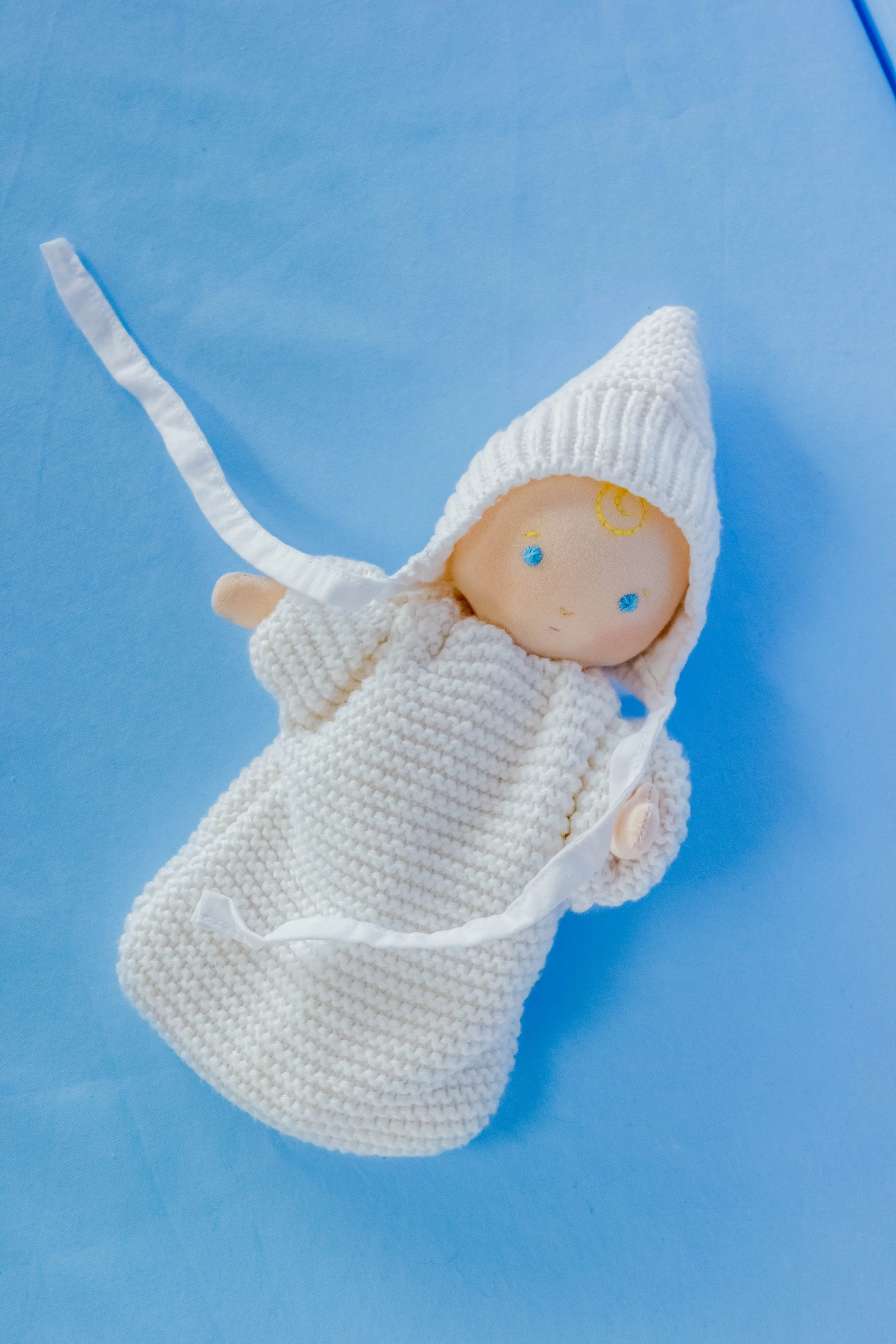 A handmade doll with a crocheted white outfit and a white knit hat with a point, lying on a blue background.