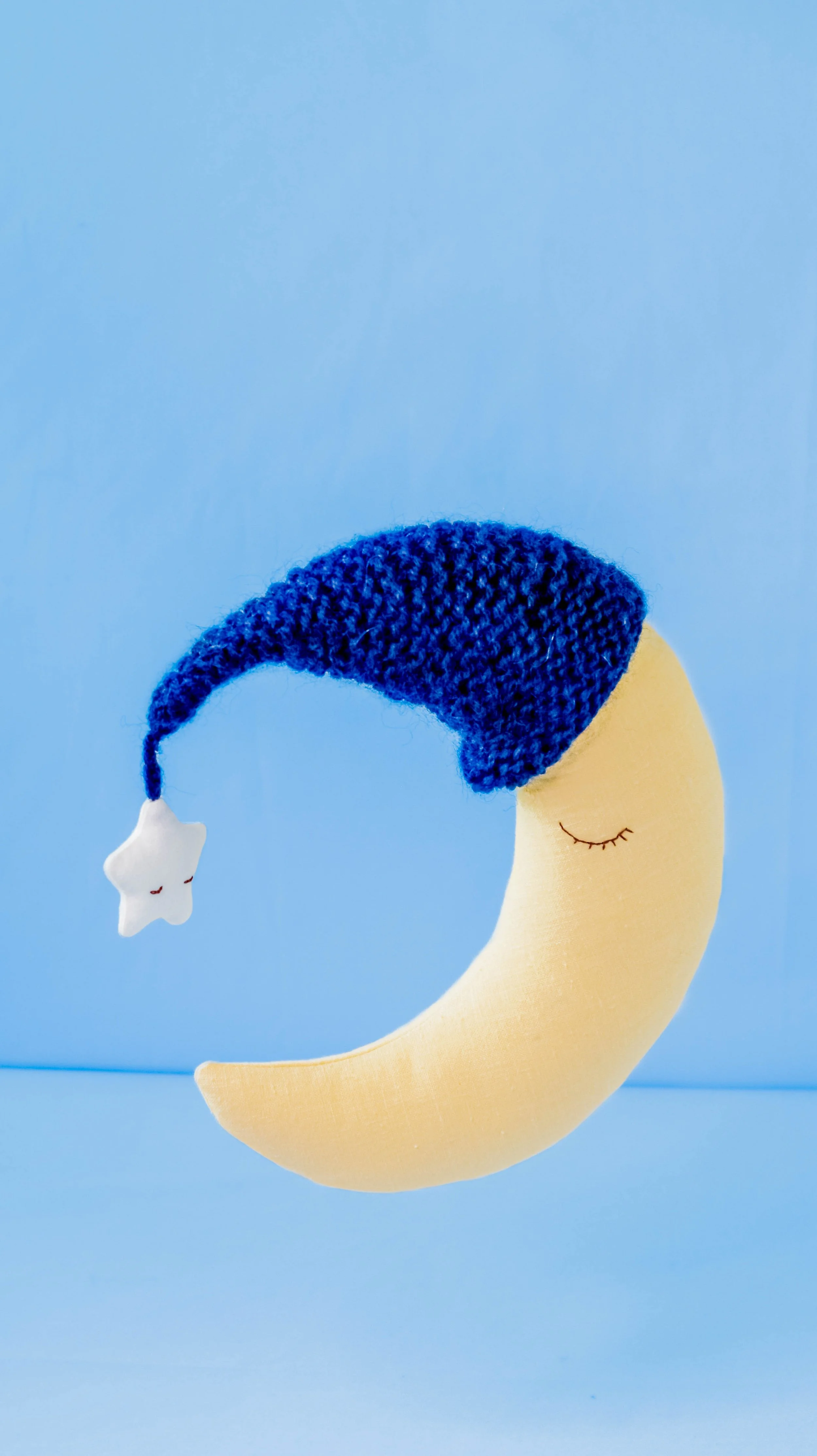 Fabric moon with closed eyes, wearing a blue knitted nightcap with a star hanging from the tip, set against a light blue background.