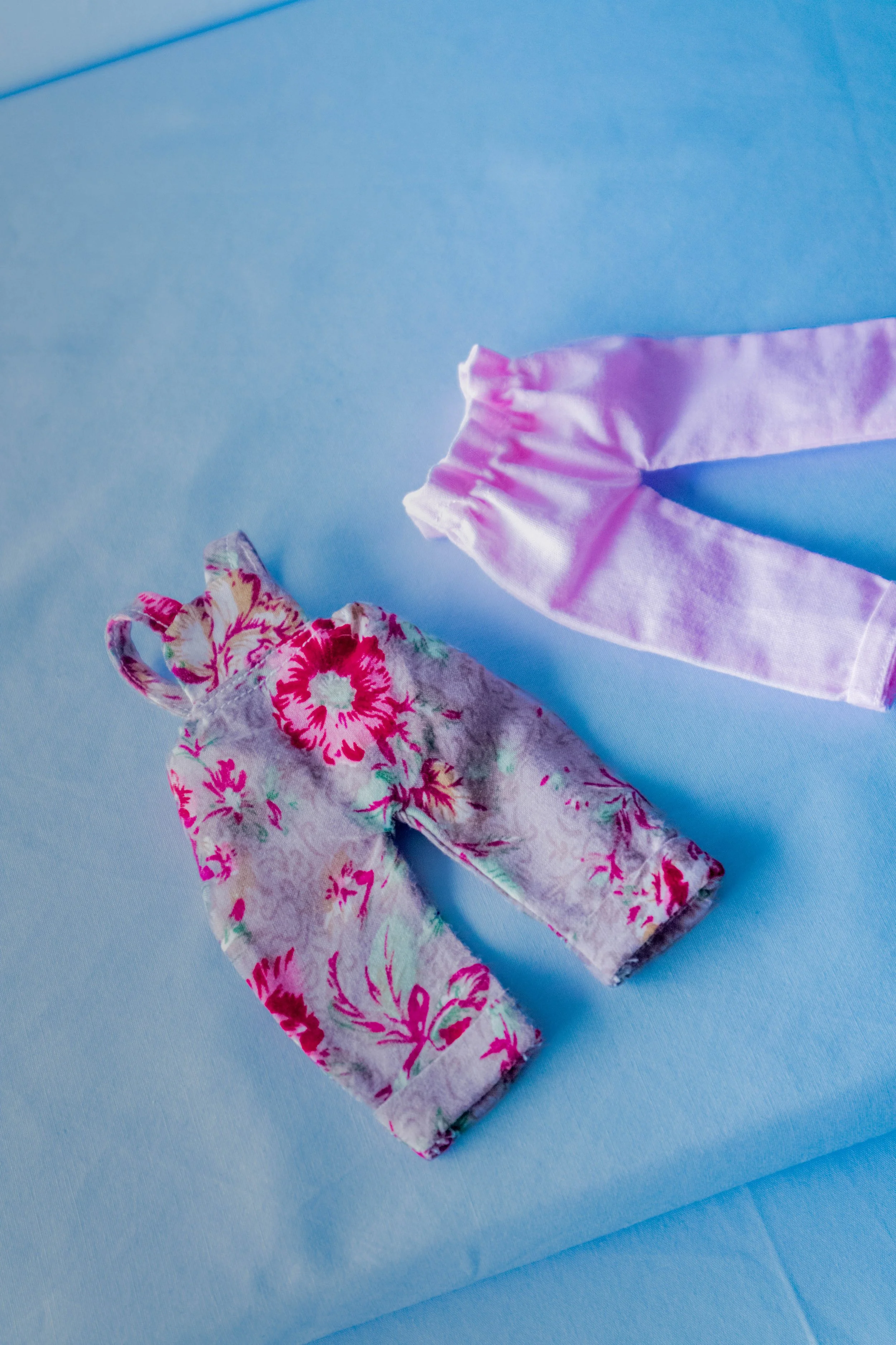 Two fabric face masks, one with pink and red floral pattern and the other with light purple with white edge detail, placed on a light blue surface.