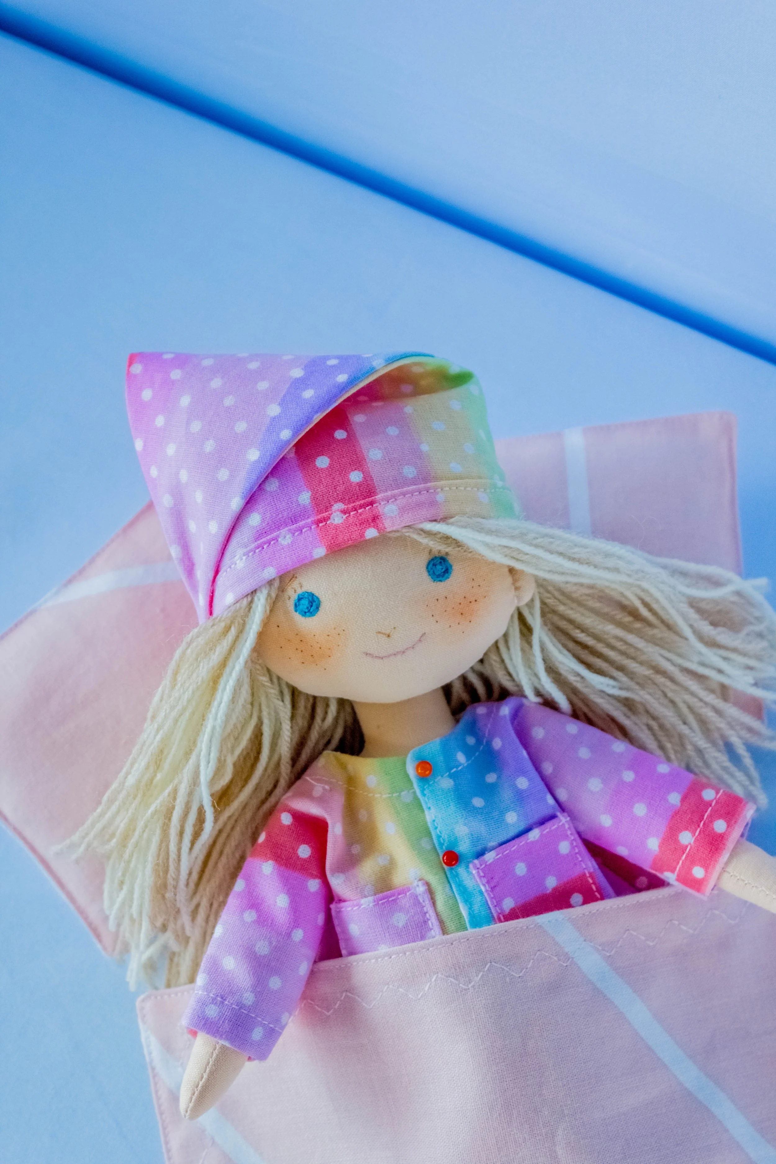 Close-up of a doll with blonde hair, wearing a pink polka-dotted hat and rainbow-colored outfit, lying on a pink pillow with a blue background.