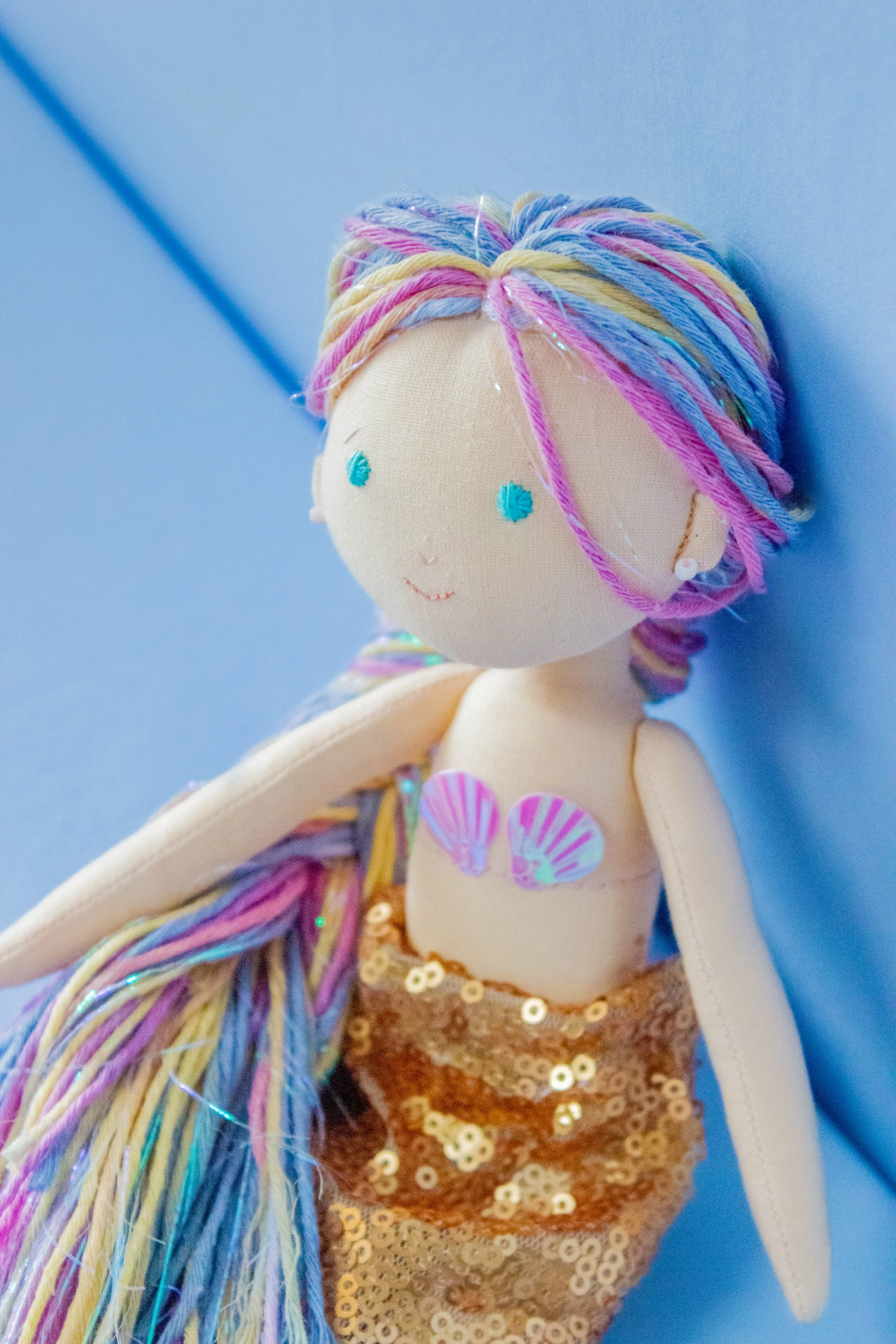 Close-up of a mermaid doll with a smiling face, colorful yarn hair, a sequined tail, and seashell details, against a blue background.