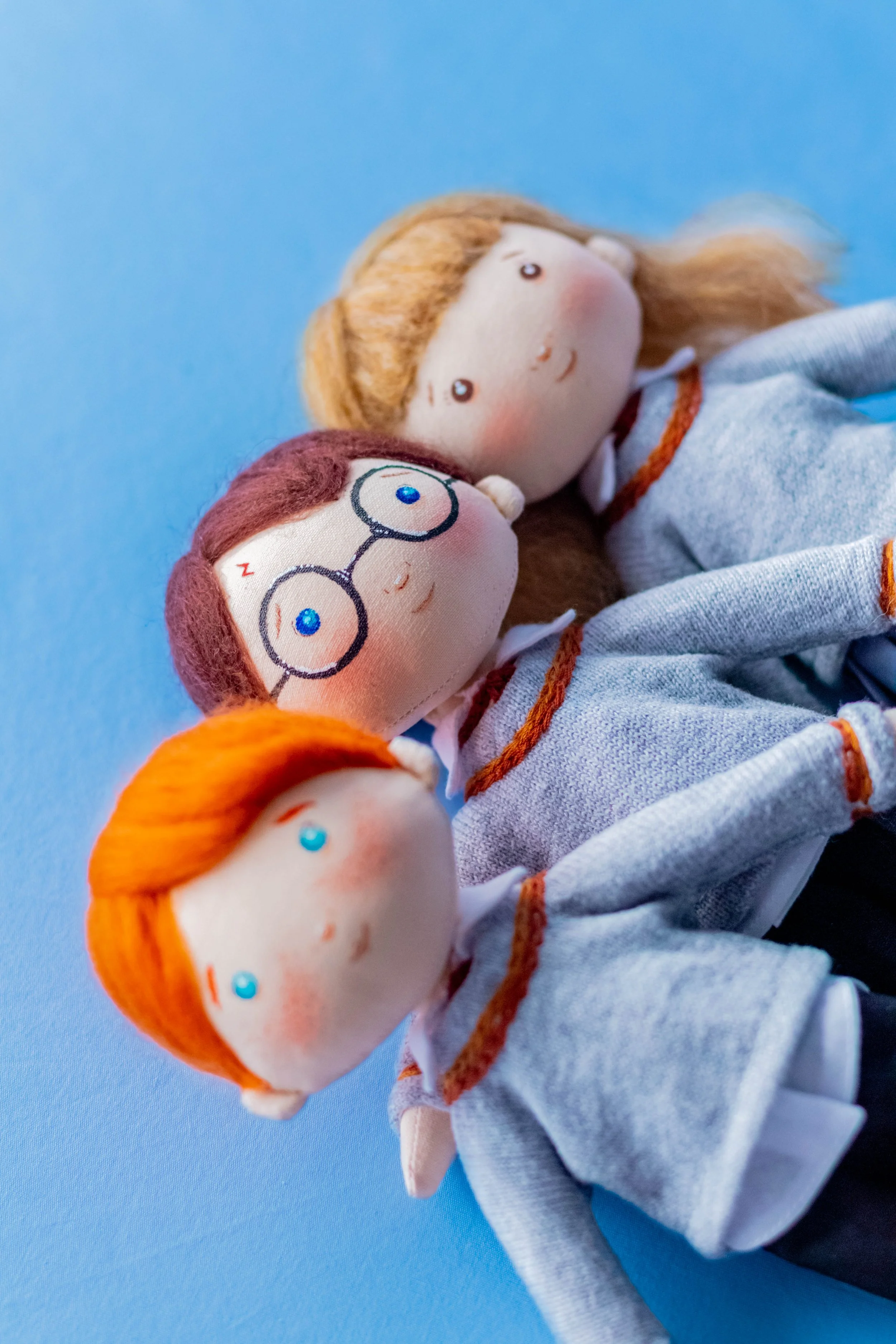 Three plush dolls with cartoonish faces, wearing glasses, and dressed in gray and white clothing, lying on a light blue surface.