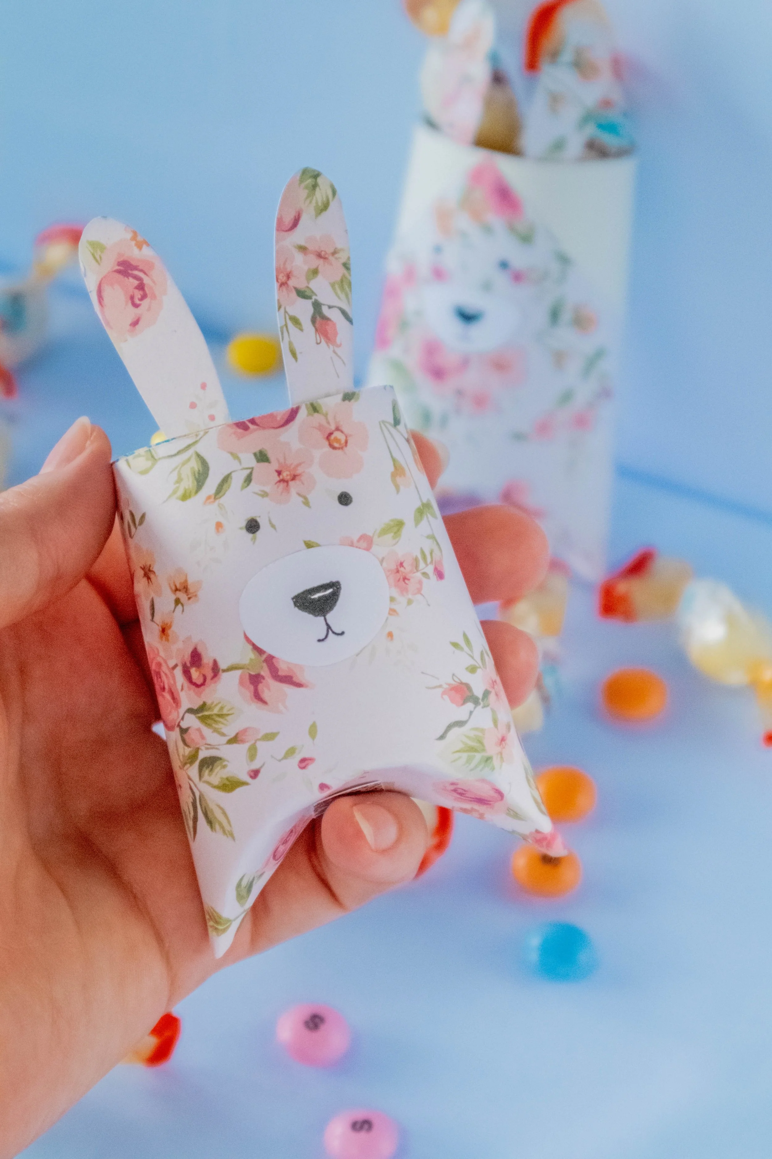 Person holding a small floral paper bag with bear illustration, with colorful candies scattered on blue surface in the background.
