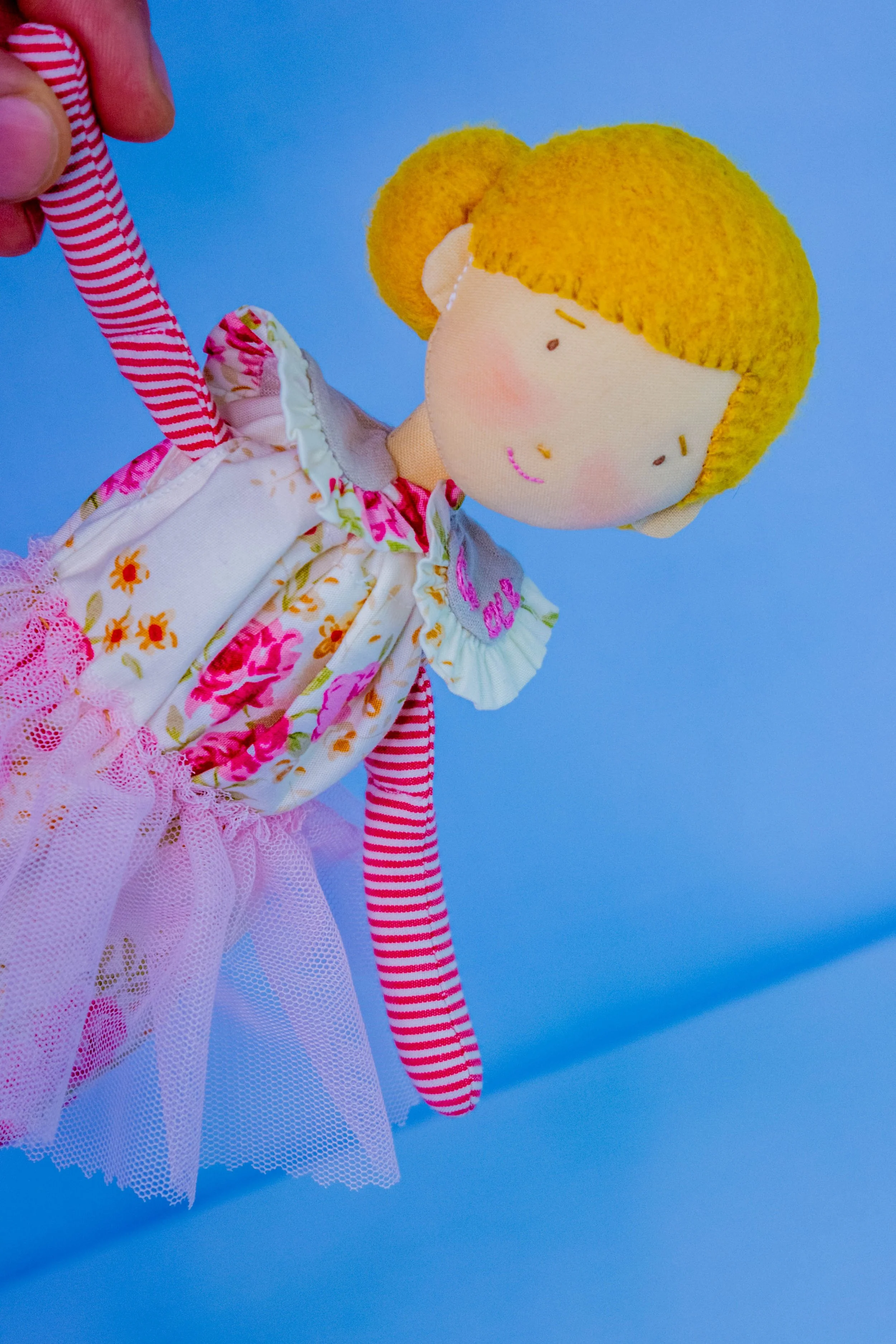 Close-up of a handmade doll with yellow yarn hair, a floral dress with pink and orange flowers, a pink tulle skirt, and striped pink and white fabric. The doll's face has simple features with a small smile, and it is set against a plain blue backgrou