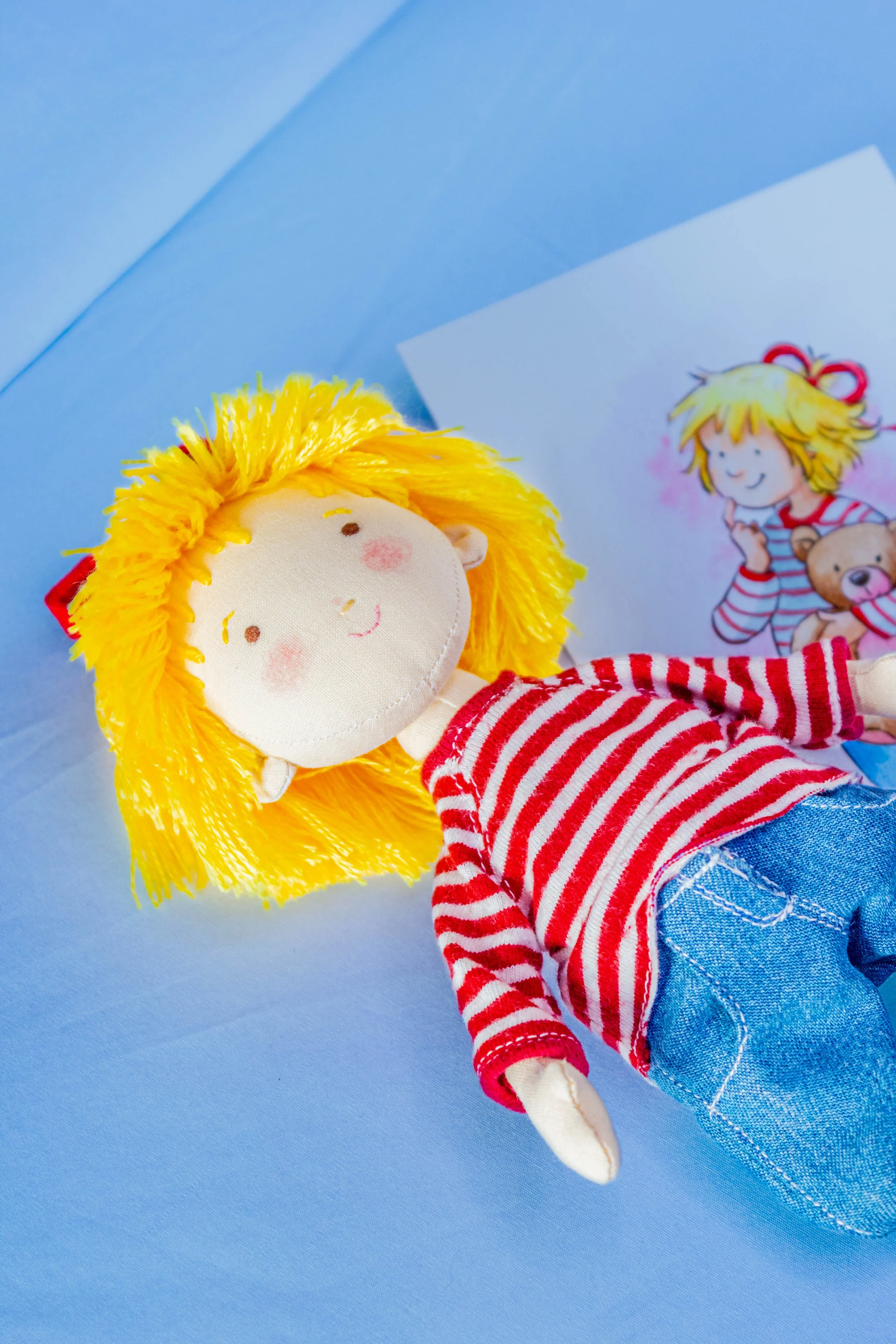 A handmade cloth doll with yellow yarn hair, wearing a red and white striped shirt and blue jeans, lying on a light blue surface next to a drawing of a girl with blonde hair, red ribbon, striped shirt, and teddy bear.