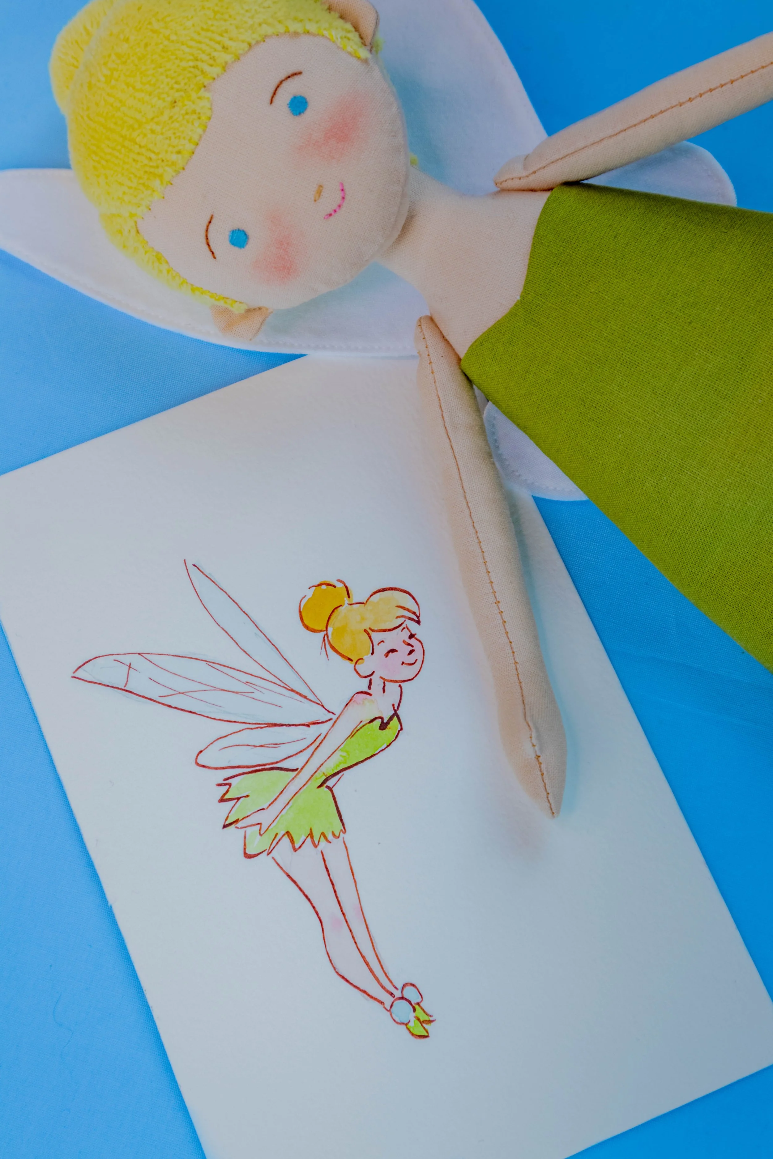 A plush doll with yellow hair, blue eyes, and pink cheeks lying on a blue surface. Beside it is an open sketchbook with a colorful drawing of a fairy with yellow hair, pink cheeks, and green dress, and purple wings.