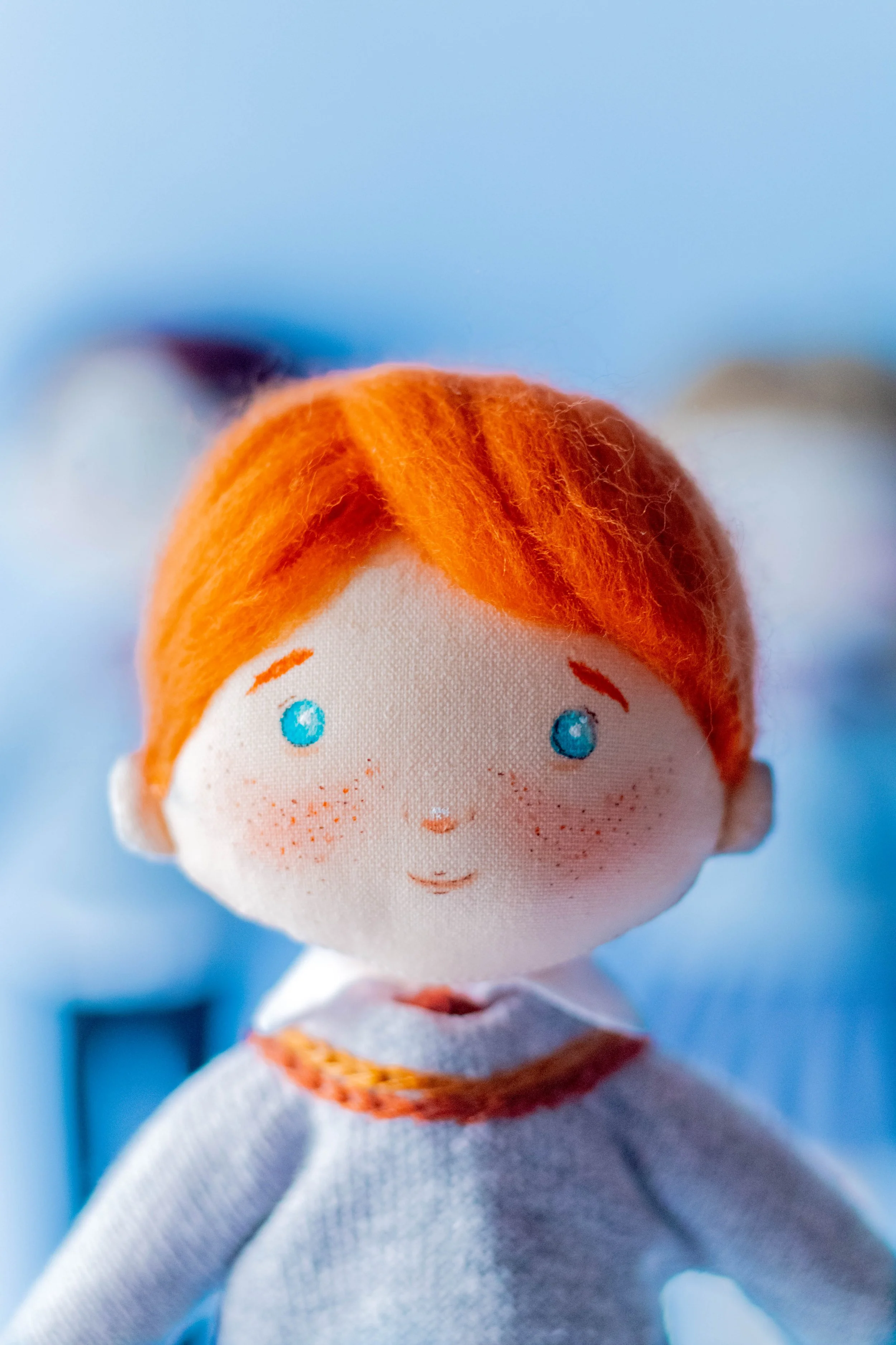 Close-up of a plush doll with orange hair, blue eyes, freckles, and a white outfit, set against a blue background.