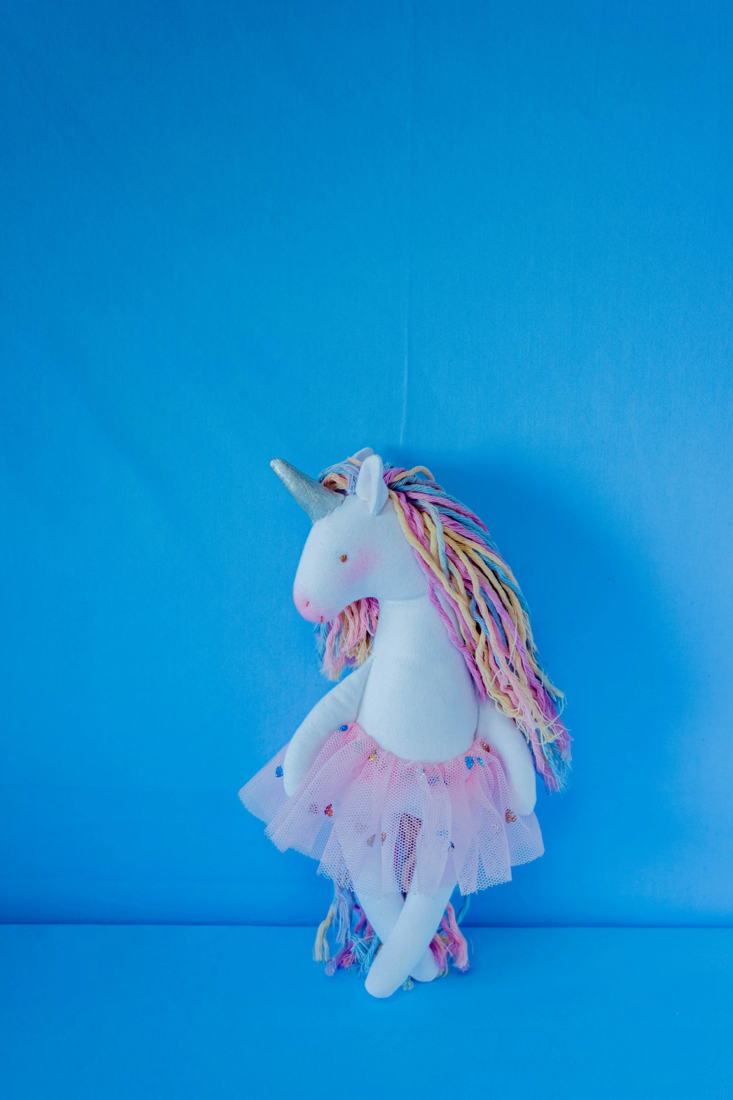 A plush white unicorn stuffed animal dressed as a ballerina with a pink tutu, multicolored yarn hair, and a silver horn, posed on a bright blue background.