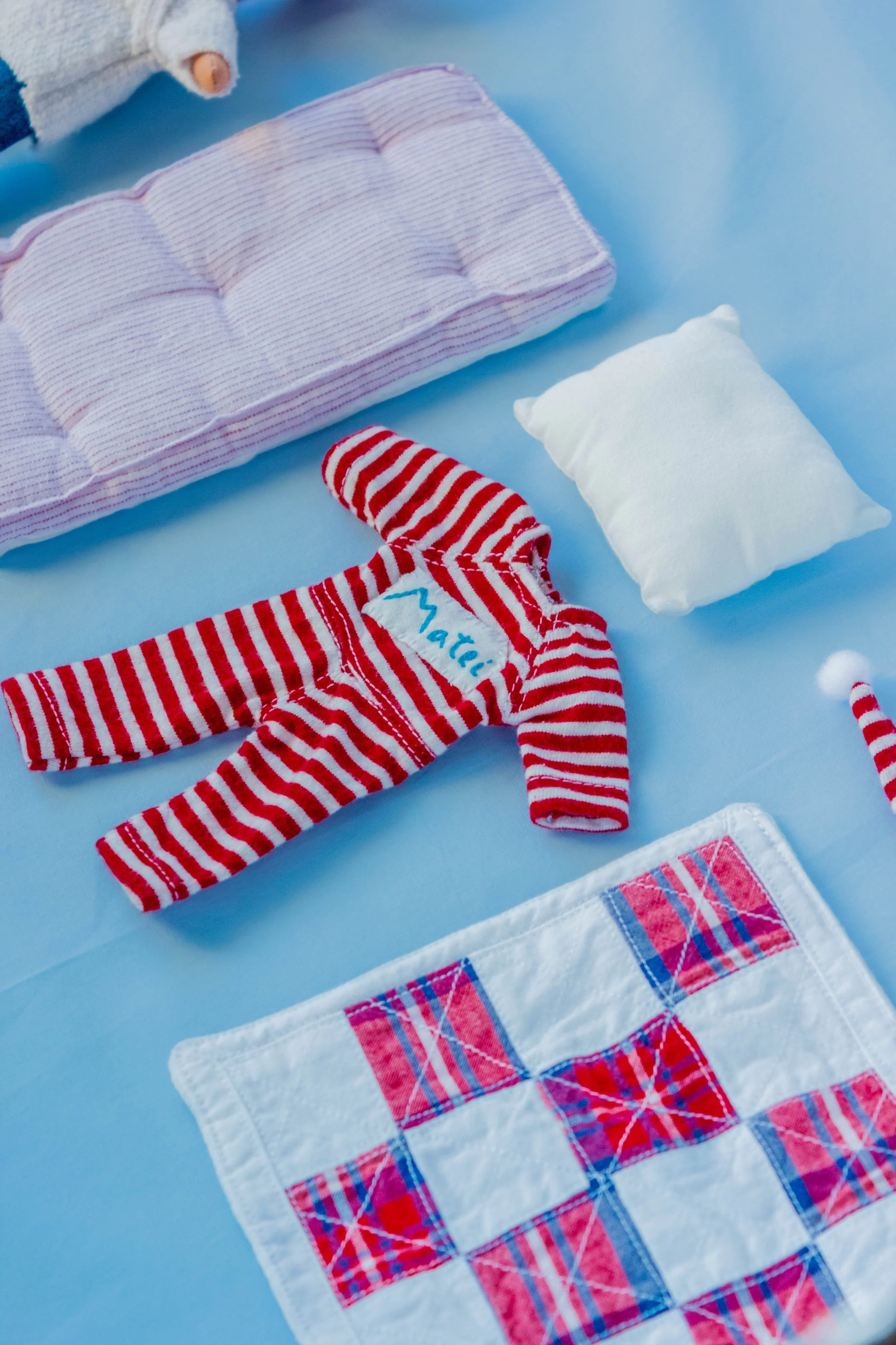 Close-up of various fabric items with red and green plaid patterns on a light blue background, including a striped sleep mask, a white fabric with pink plaid trim, and a small pillow.