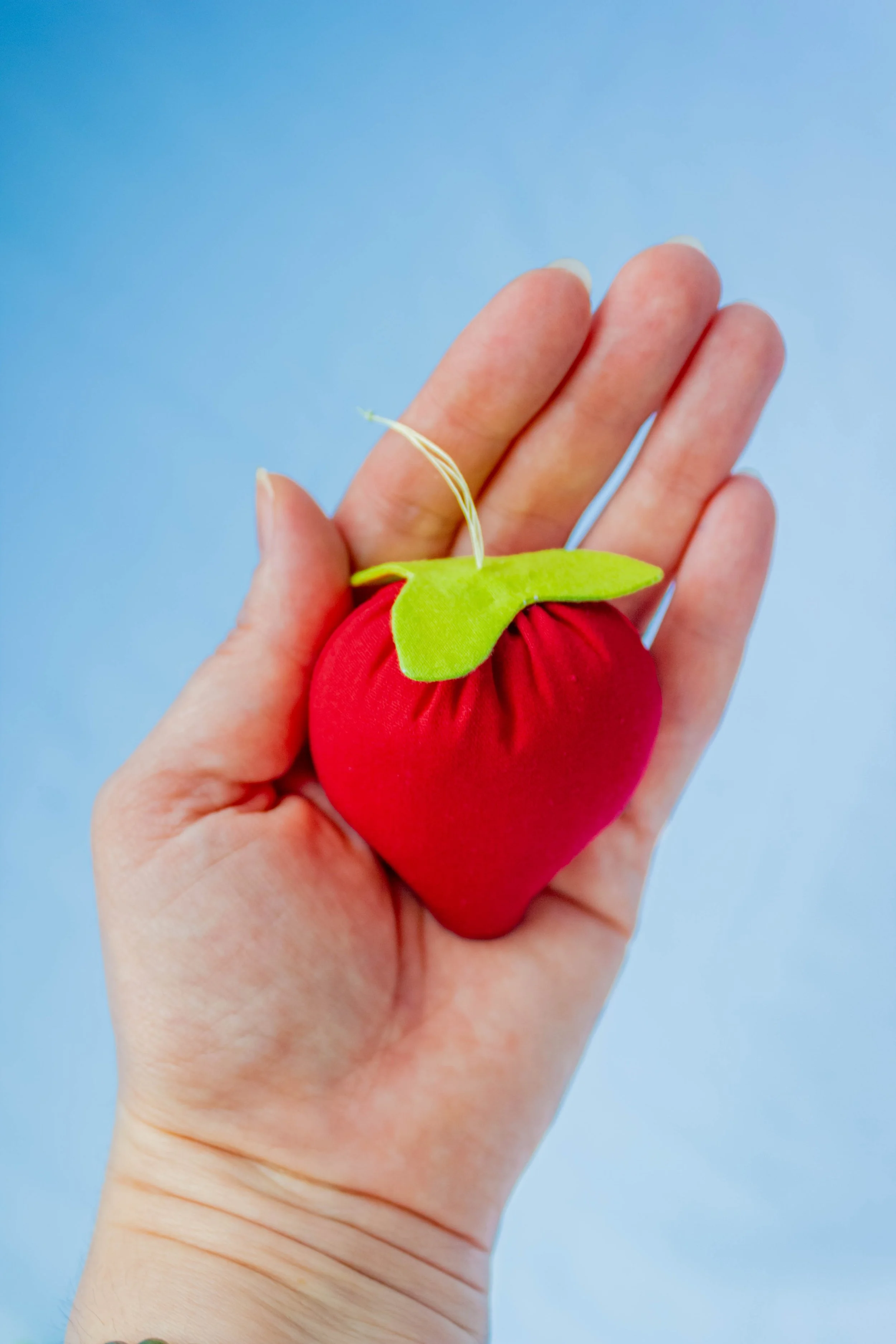 A hand holding a fabric strawberry ornament with a green leafy top and a string for hanging, against a light blue background.