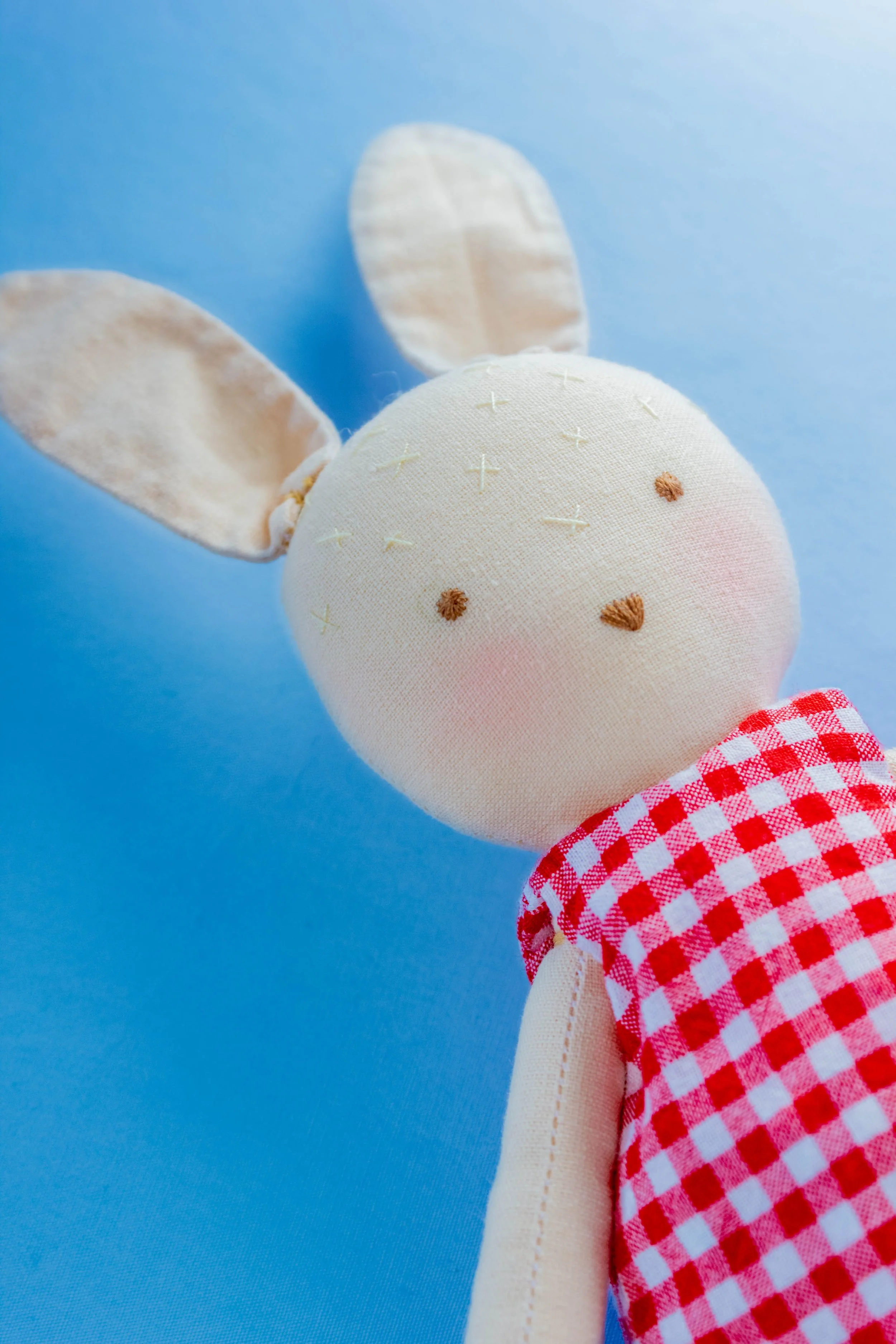 Close-up of a handmade fabric bunny toy with a beige face, embroidered features, and large ears, wearing a red and white checkered dress, against a solid blue background.