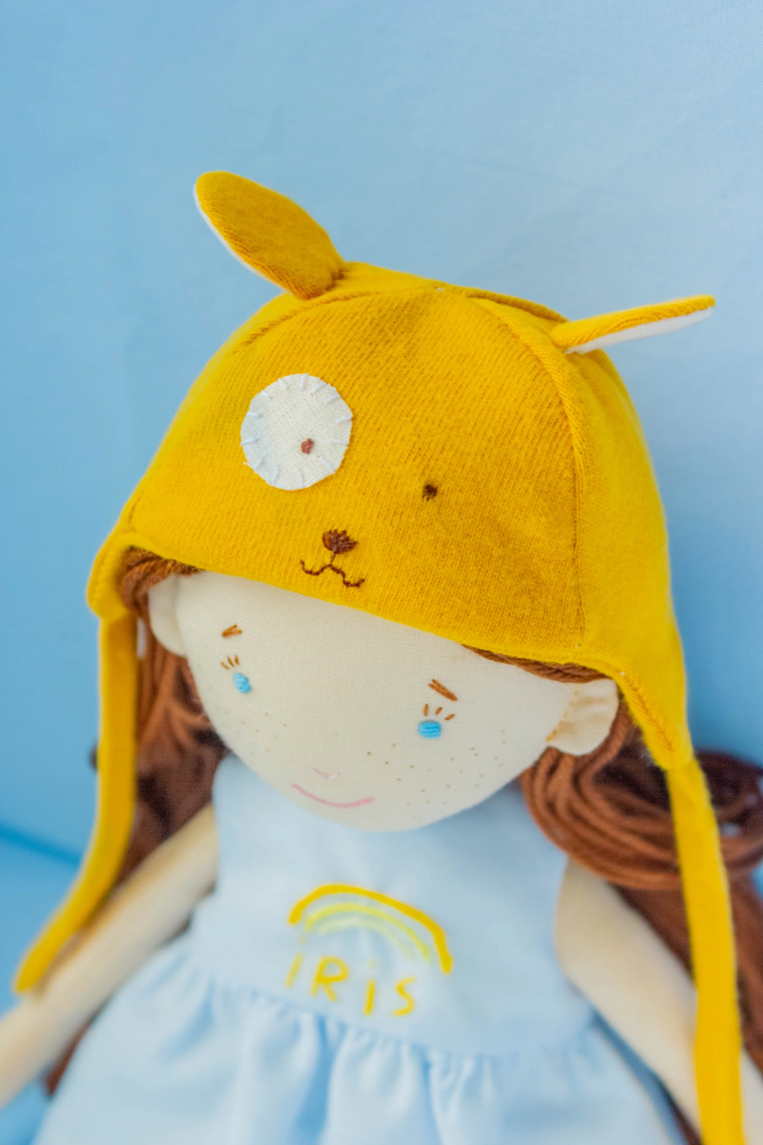 Close-up of a plush toy girl with brown hair, wearing a yellow hat with a bunny face, ears, and a white shirt with yellow embroidery, against a blue background.