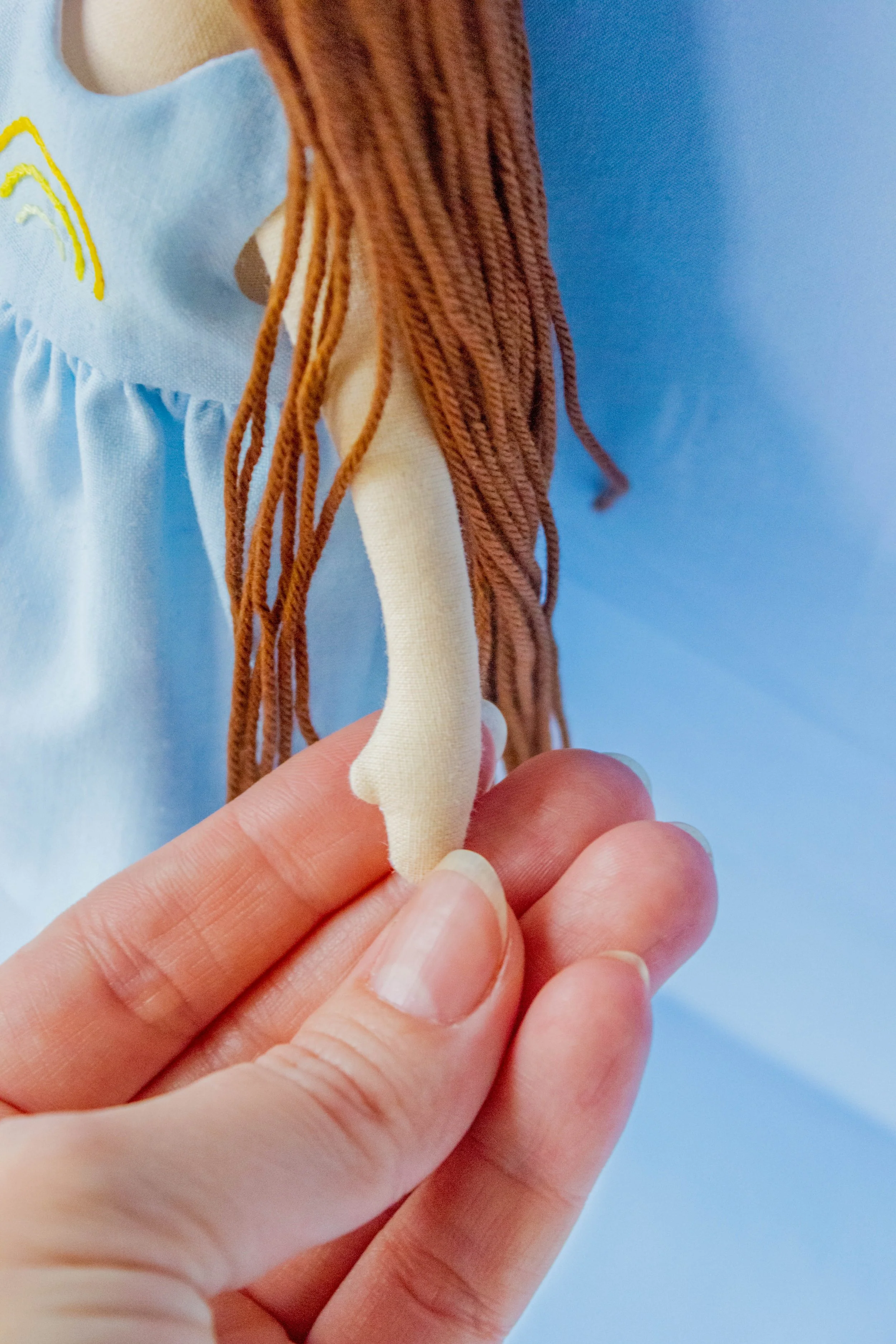 Close-up of a hand holding a small fabric doll with long brown yarn hair and a light-colored dress with yellow embroidery, against a blue background.