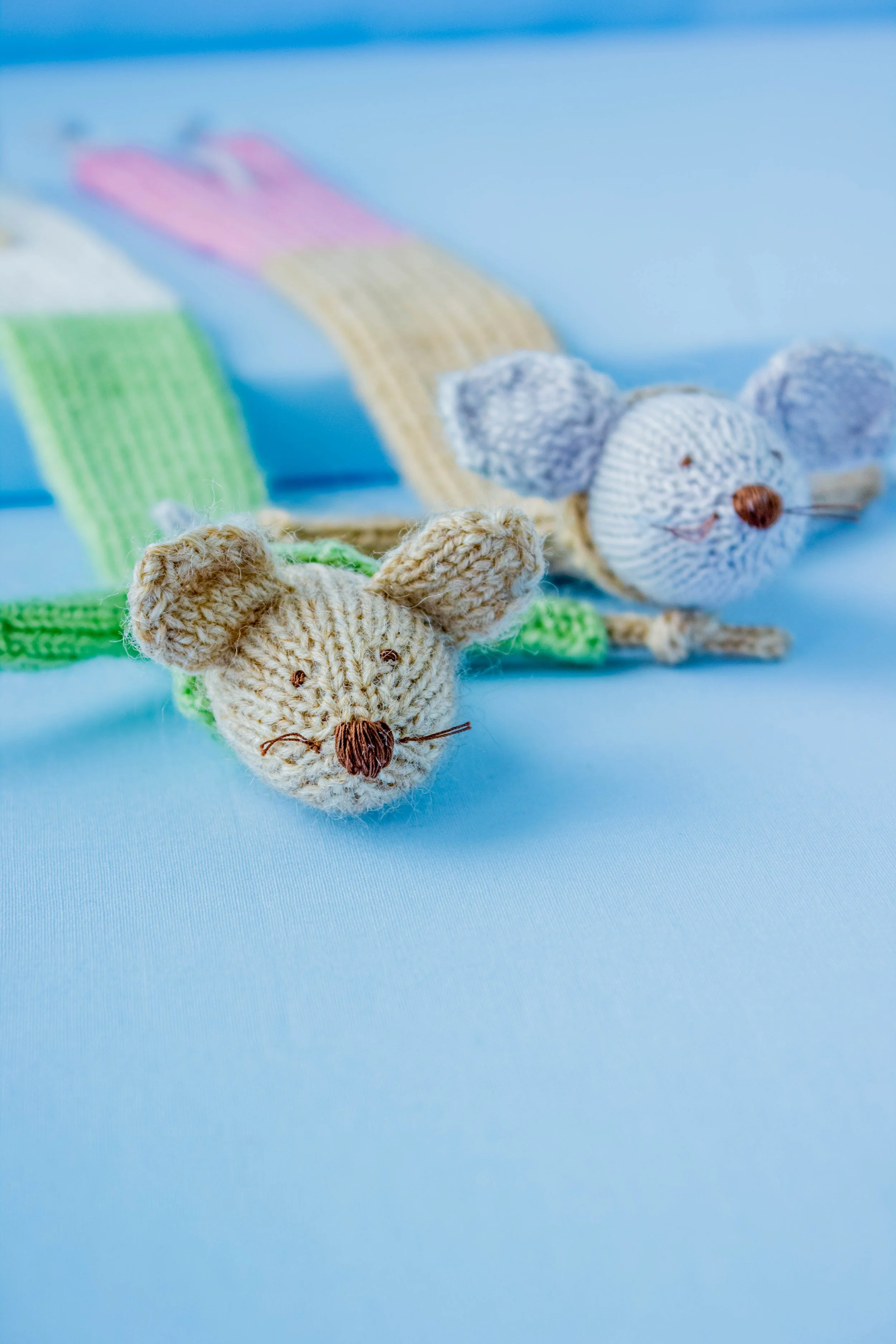 Close-up of two tiny knitted mice with a painted toy mouse in the background, against a light blue background.
