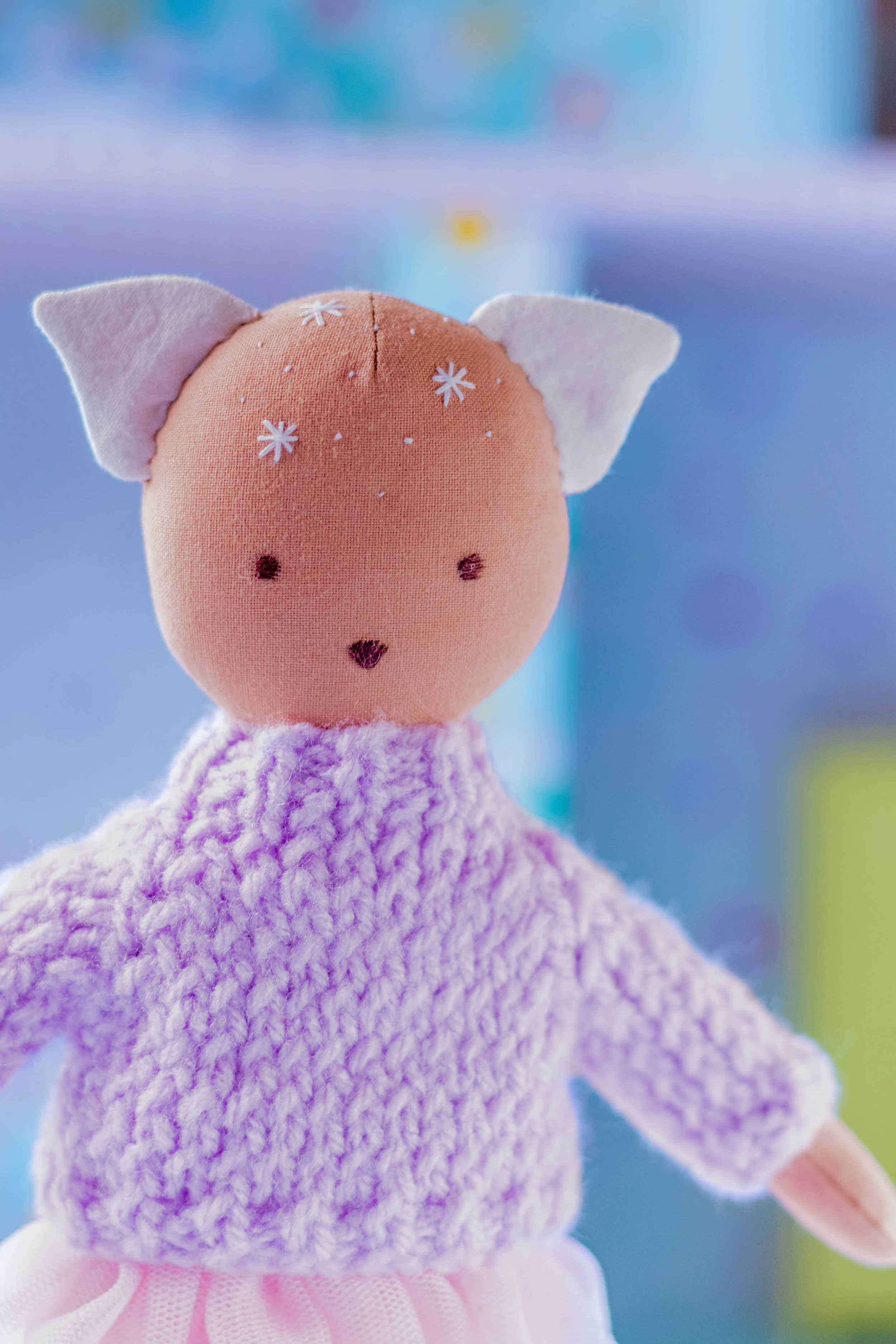 A close-up of a handmade plush toy resembling a cute animal, with a pink face, small dark eyes, a tiny nose, and a wide, fluffy purple body. The toy has white felt ears with pink inner details and is wearing a ruffled pink skirt. The background is bl