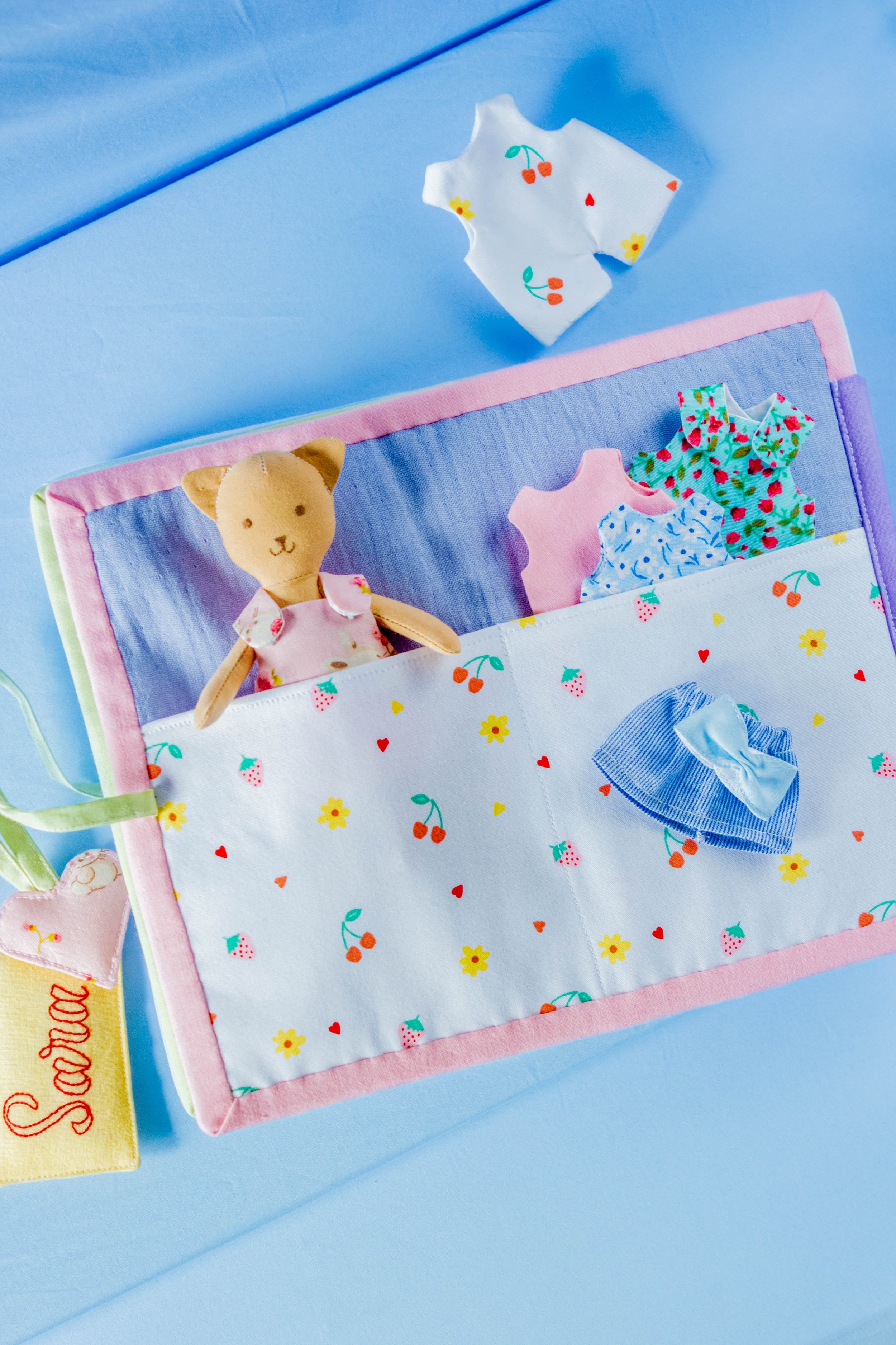 Open fabric book with clothing and a stuffed animal on a blue surface.