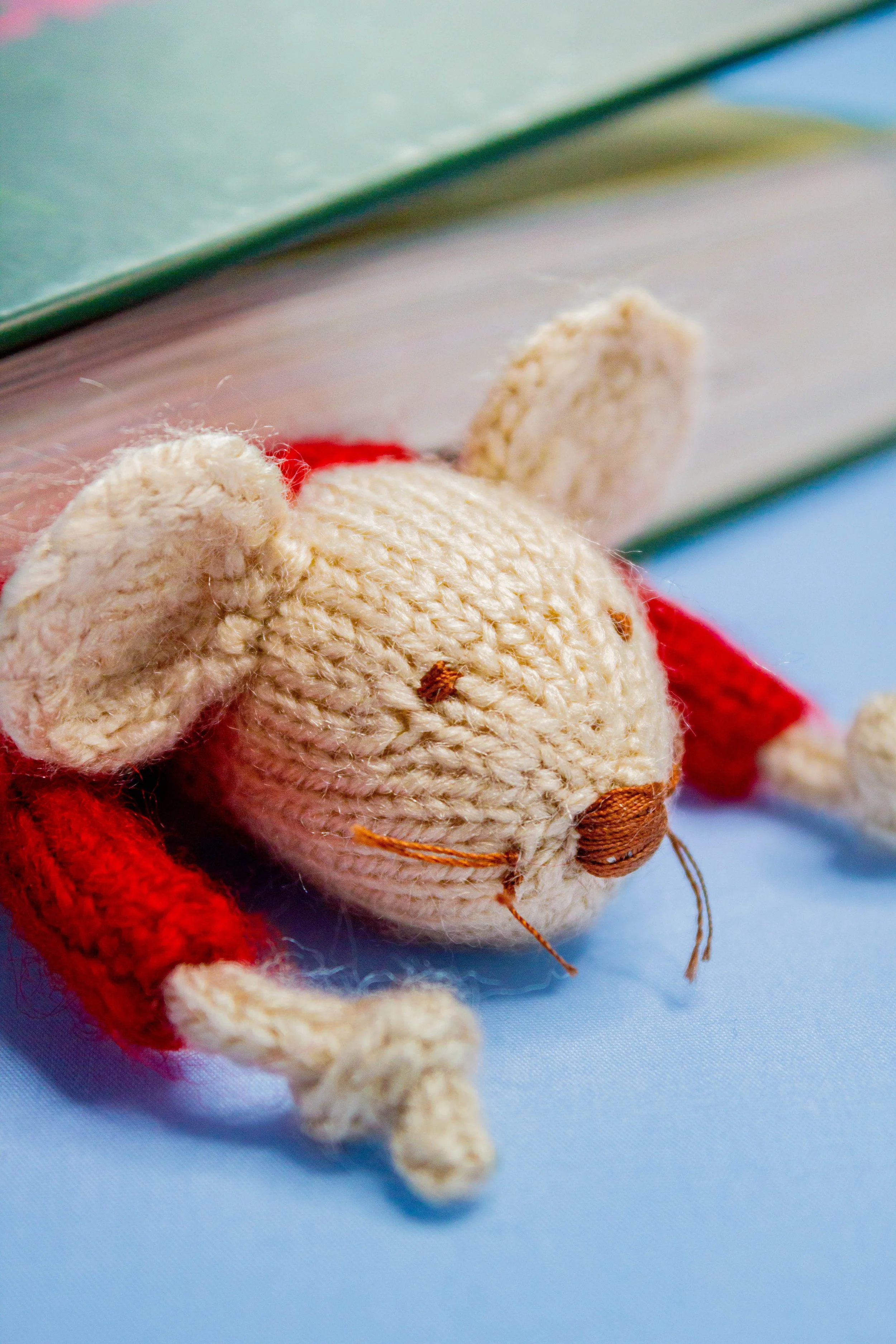 Close-up of a plush toy mouse with a red body, cream-colored face, and large round nose, lying on a light blue surface with a book in the background.