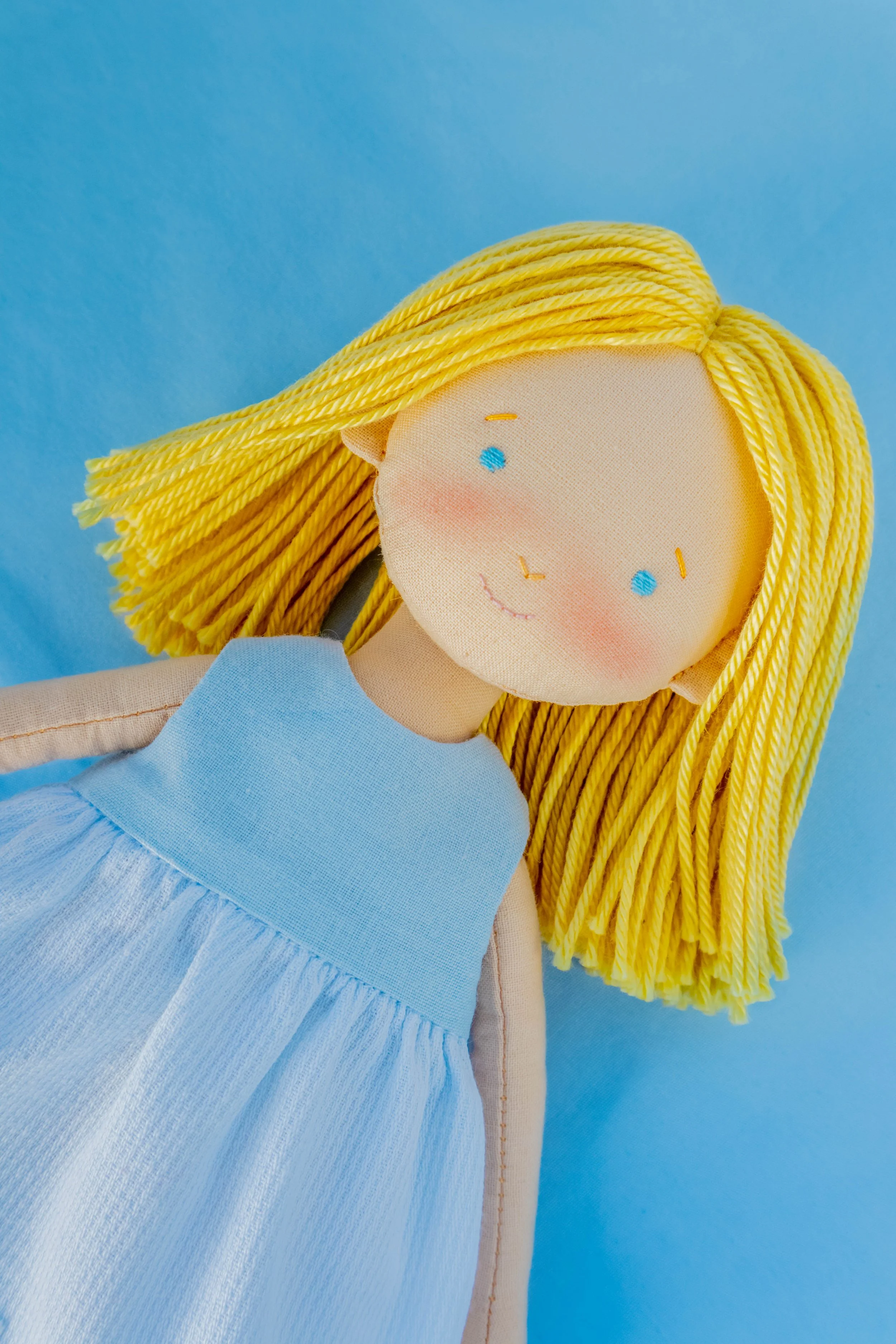A handmade fabric doll with yellow yarn hair, blue dress, and embroidered face, set against a light blue background.