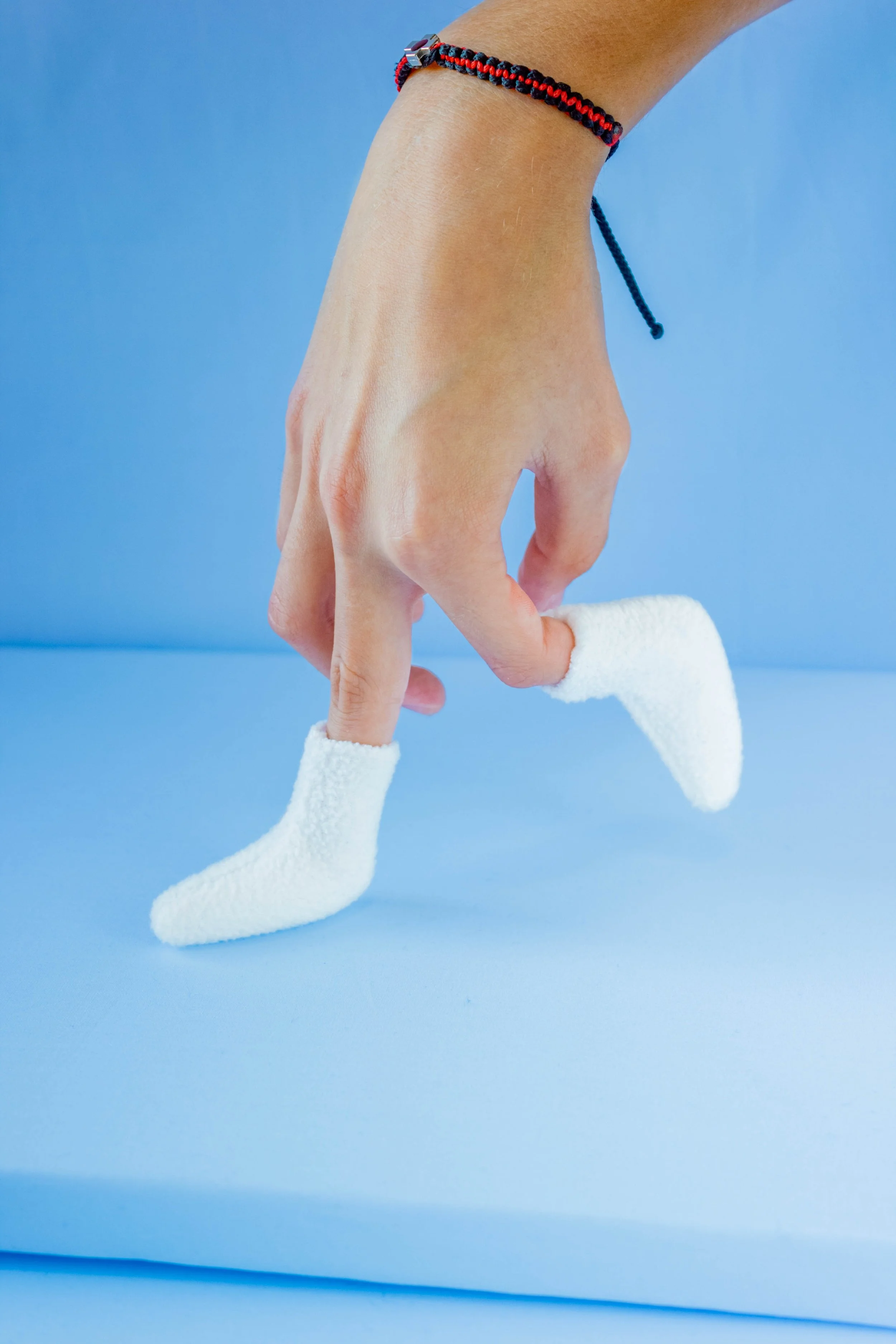 A human hand, supposedly a mannequin, is dressed in a white sock, with fingers relaxed against a light blue background. The hand is wearing a piece of jewelry on the wrist, a woven bracelet with red, black, and blue strands.