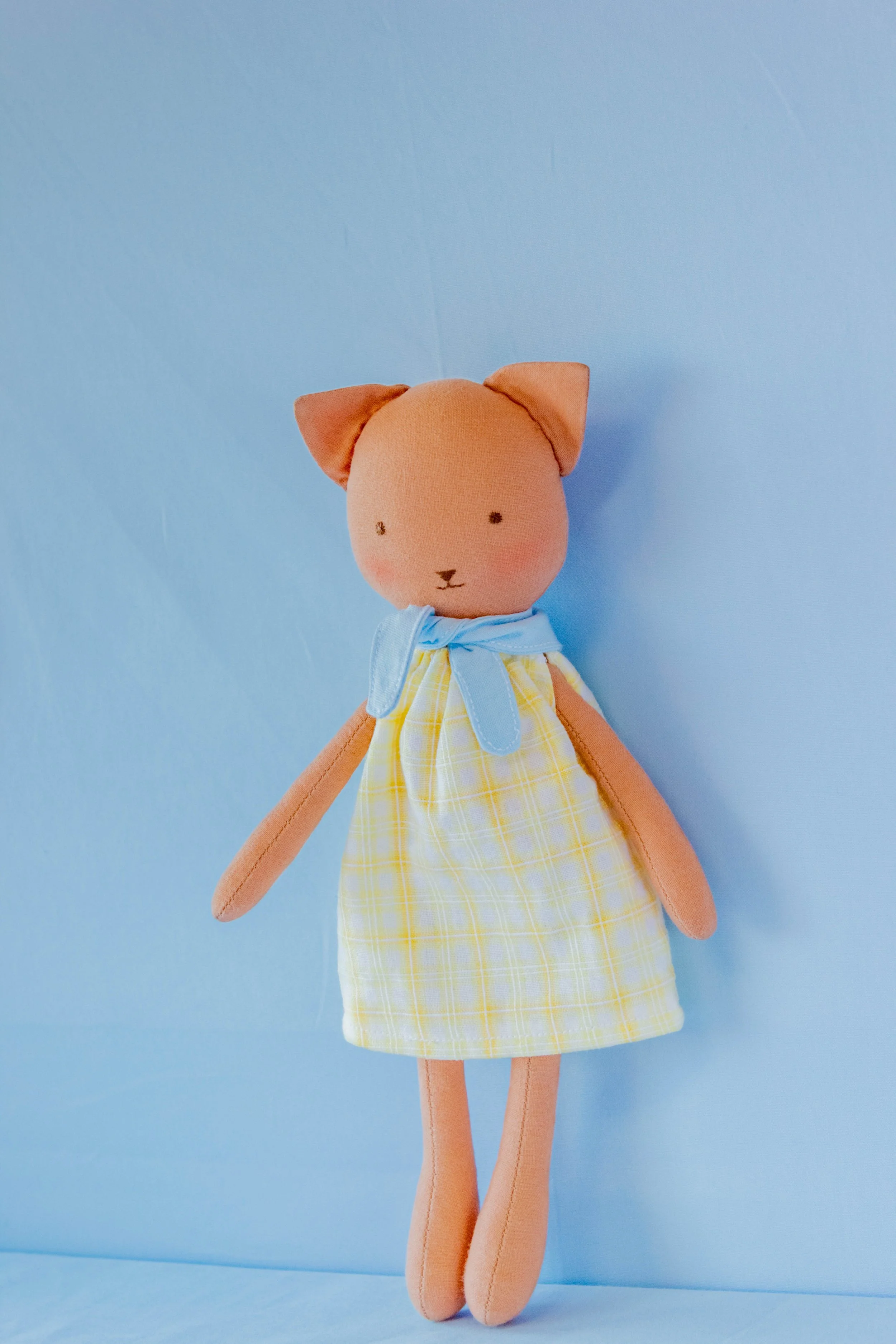 A stuffed animal resembling a cat with an orange body, sitting against a blue background. It is wearing a yellow checkered dress with a light blue bow around its neck and has ears, small embroidered eyes, and a nose.