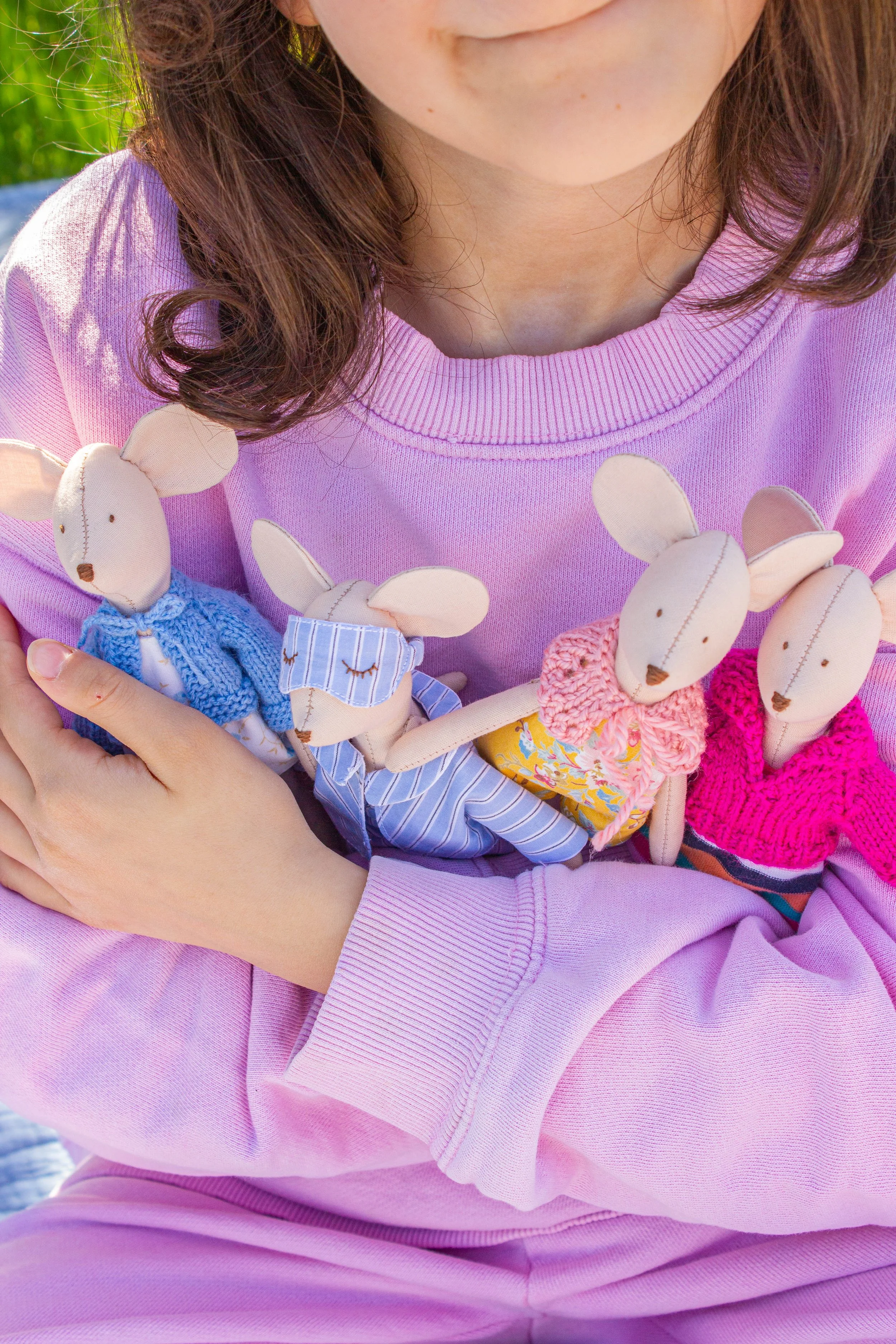 Close-up of a person with brown hair wearing a pink sweatshirt, holding several handmade stuffed mice made of fabric and yarn, with different colored clothes and accessories.