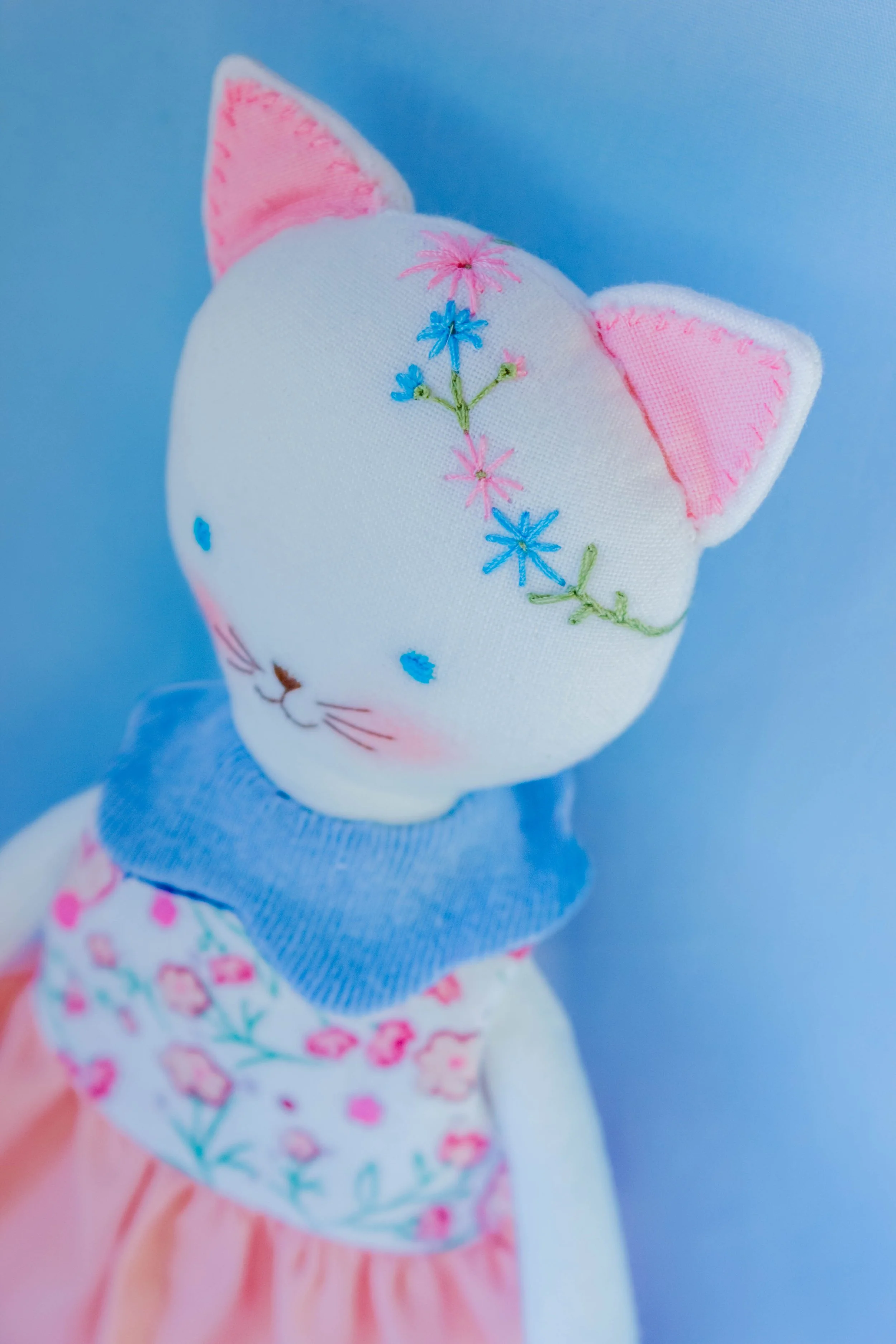Close-up of a plush toy with embroidered flowers on its white face, pink ears, a blue sweater, and pink floral skirt, against a blue background.