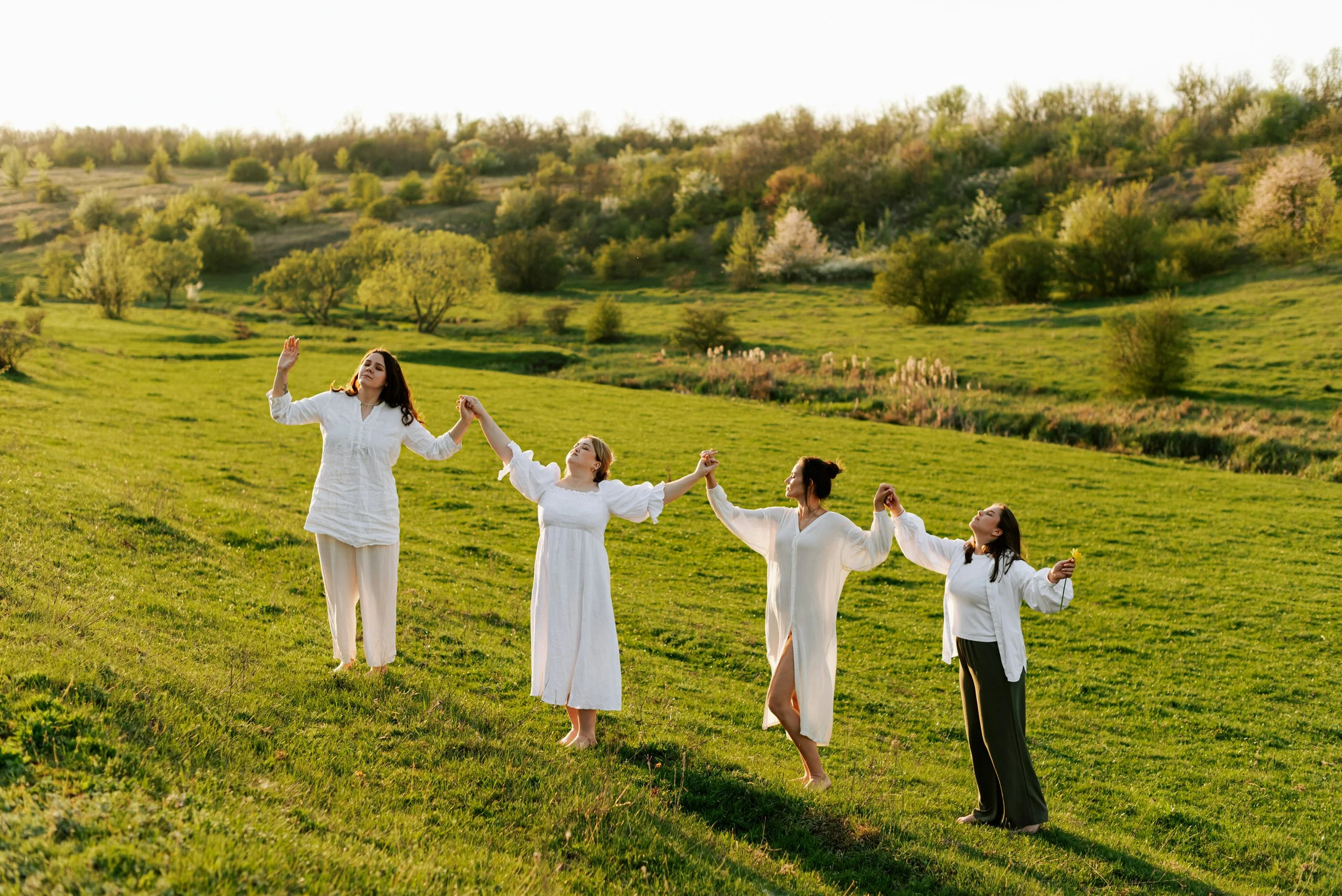 Four women in white dresses holding hands and dancing in a grassy field with trees and hills in the background during sunset.