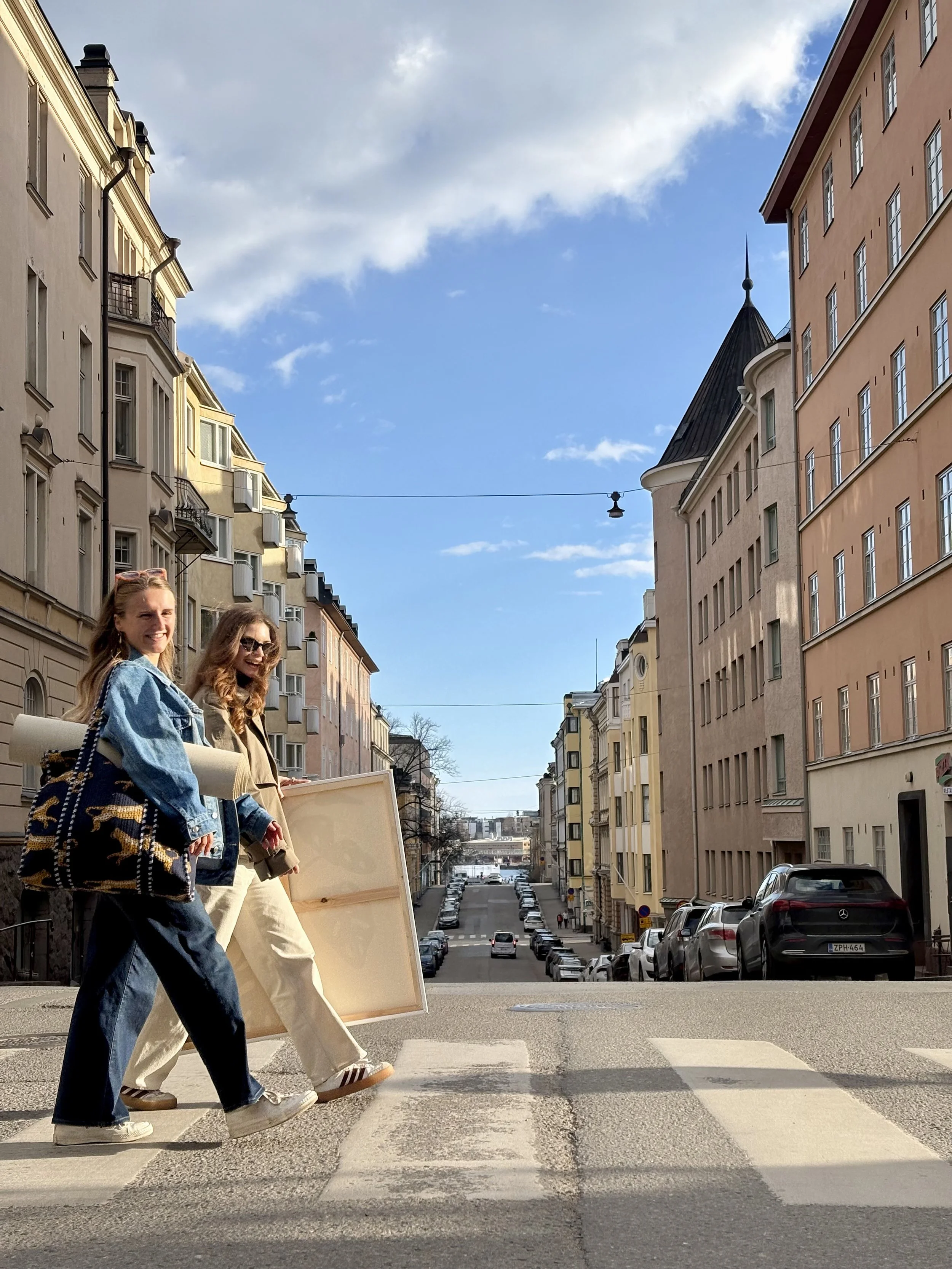 Two women walking across a street with apartment buildings on either side; one woman is holding a painting canva, and the other a yofa mat.  Both are dressed casually. Cars are parked along the street