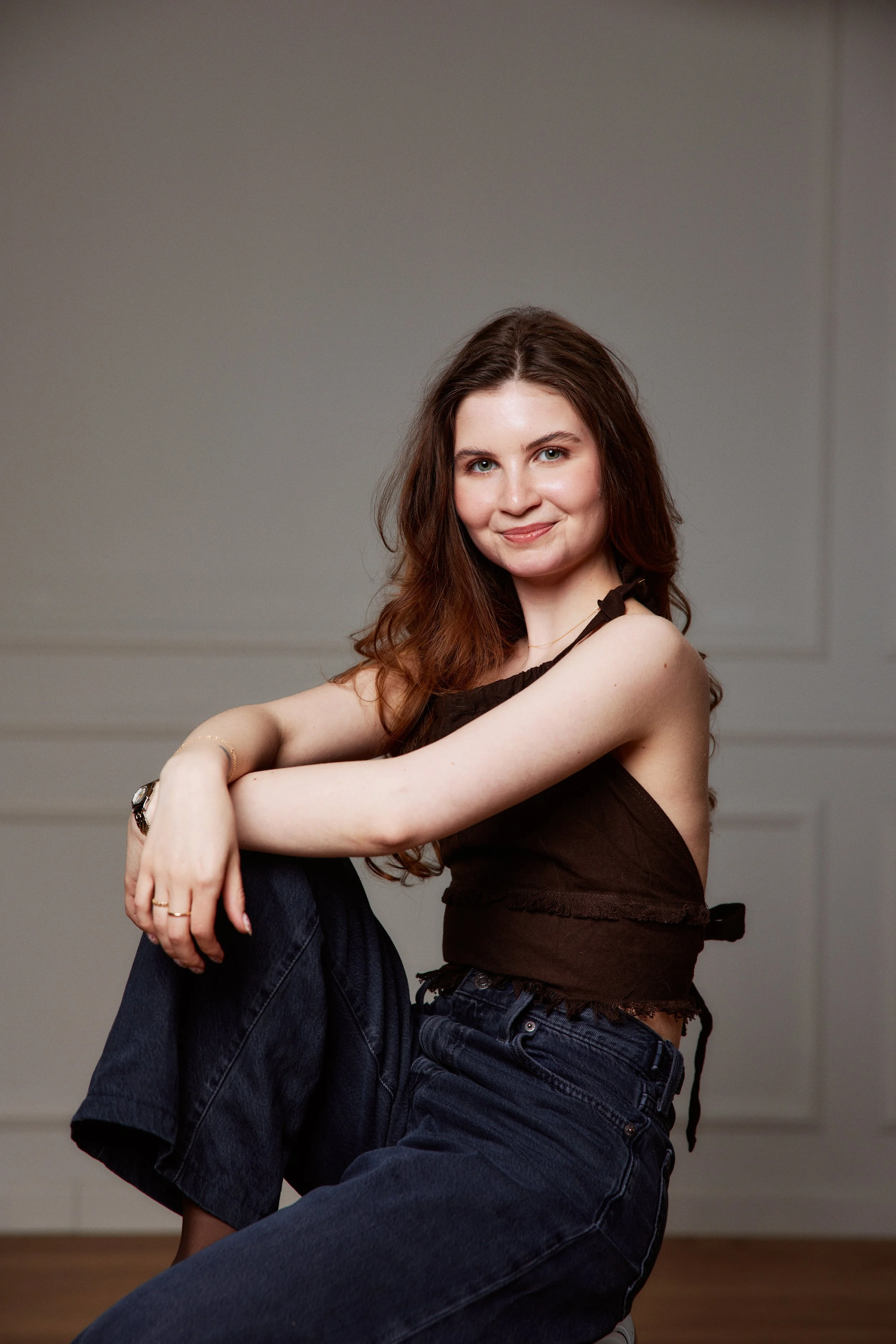 A young woman with long brown hair, wearing a brown sleeveless top and dark jeans, sitting on the floor, smiling with arms crossed over her knees, against a plain white wall background.