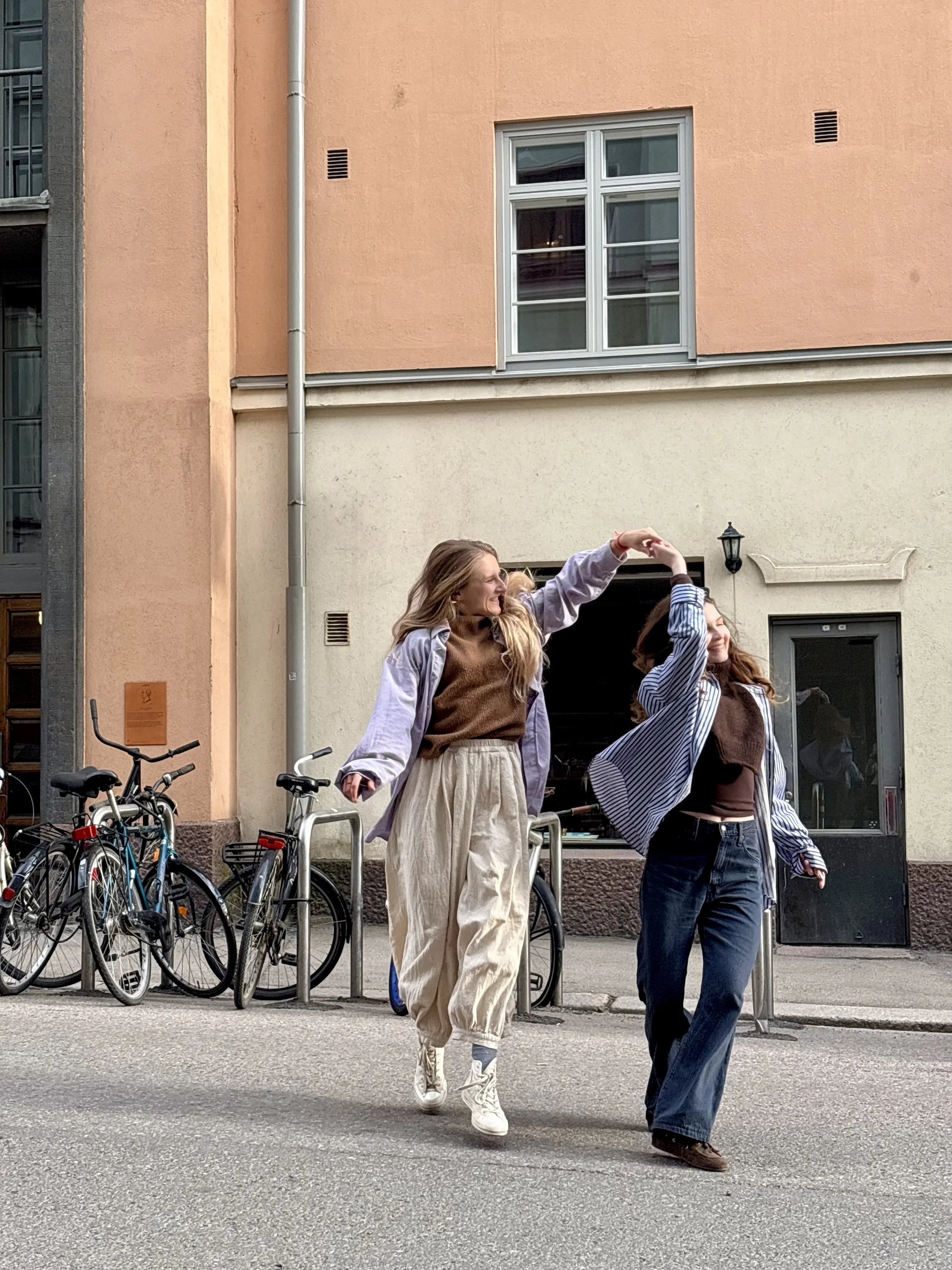 Two young women walking on the street, one with long blond hair wearing a light jacket and beige pants, and the other with brown hair wearing a striped jacket and black jeans. They are smiling and appear to be happy.