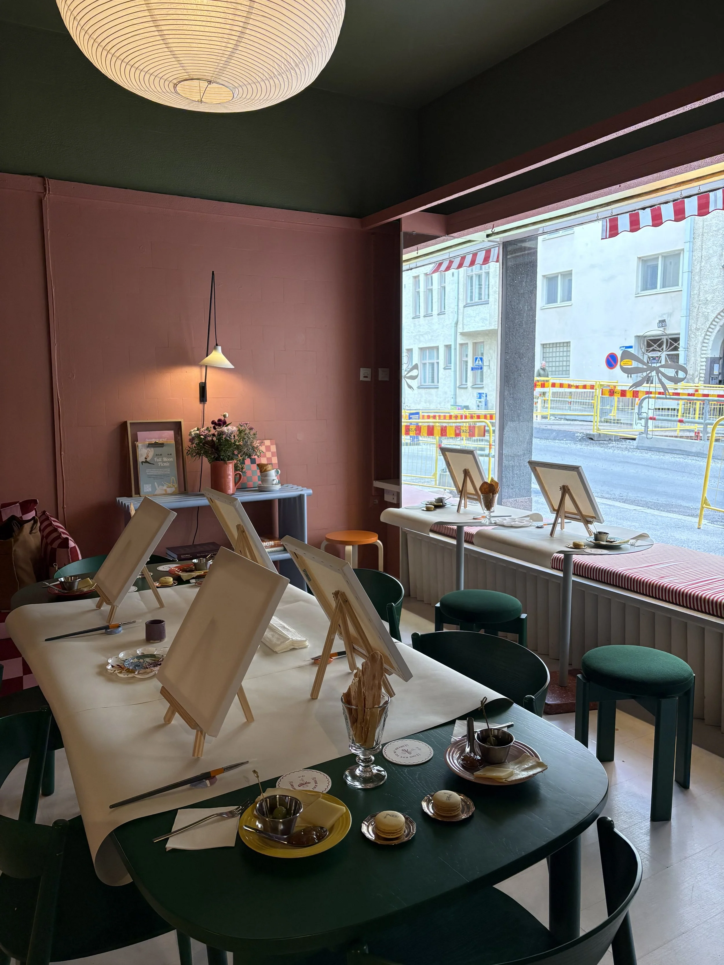 Interior of a cozy cafe or restaurant with a large window. A table set with food, including macarons, chocolates, and a glass of coffee, is in the foreground. Several easels with art are on the table, and a wall with a pinkish color and a small shelf with flowers and framed pictures is visible. The ceiling has a round paper lantern light.
