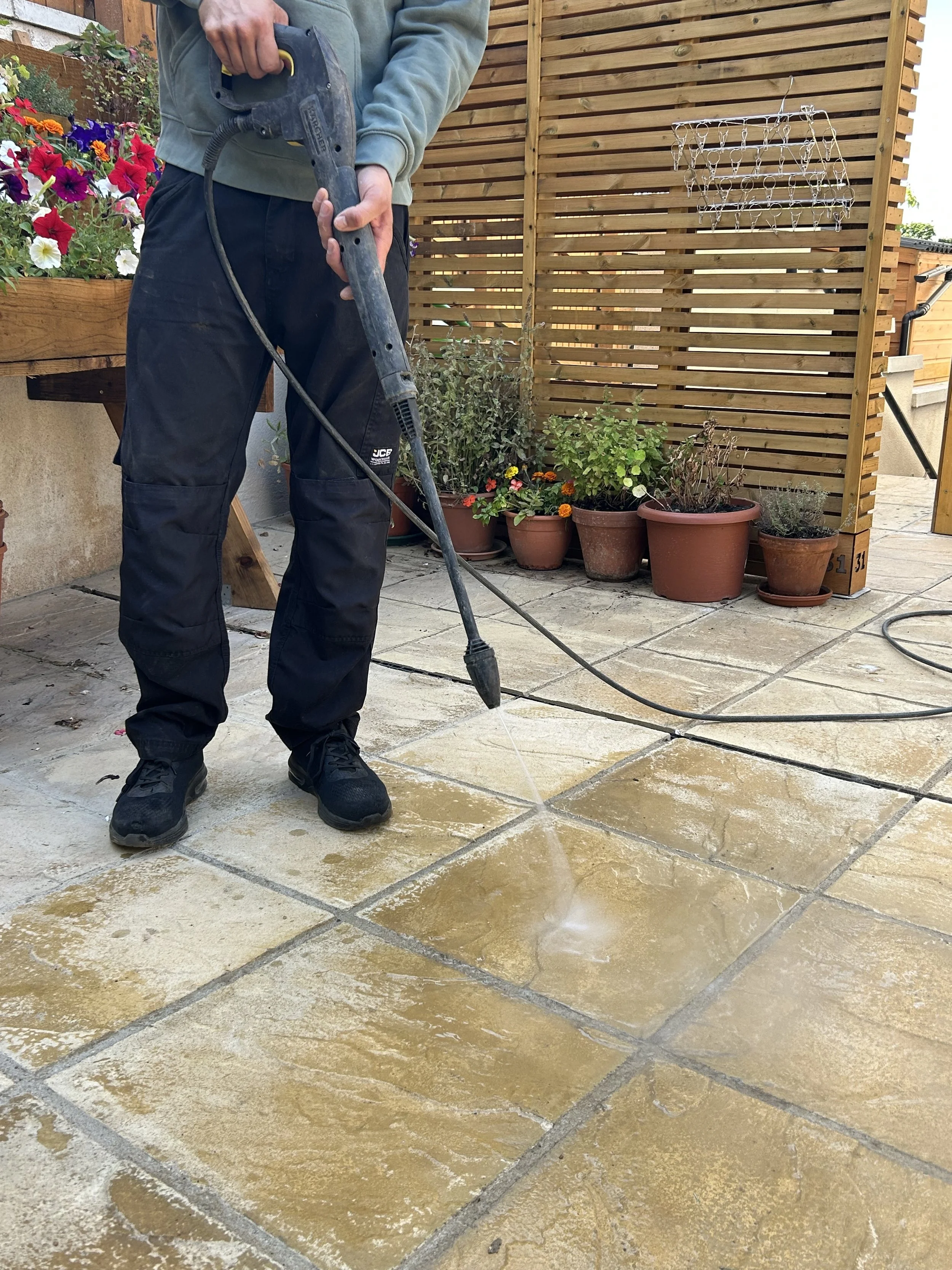 Person power washing outdoor tiled patio with potted plants and wooden fencing in background.