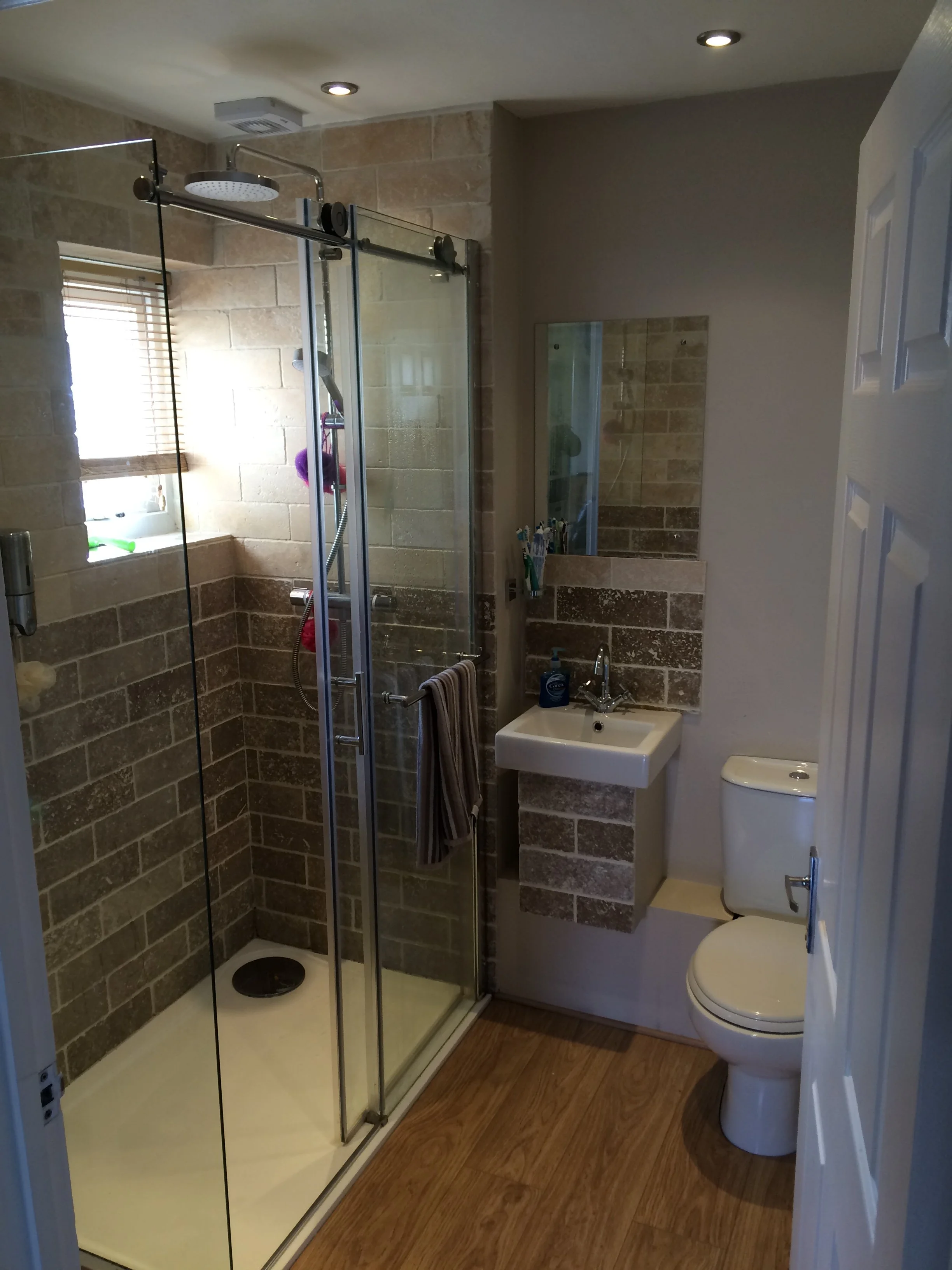 A small bathroom with a glass-enclosed shower on the left, a small wall-mounted sink with a mirror above it, and a toilet on the right; wood-look flooring and beige tiled walls.