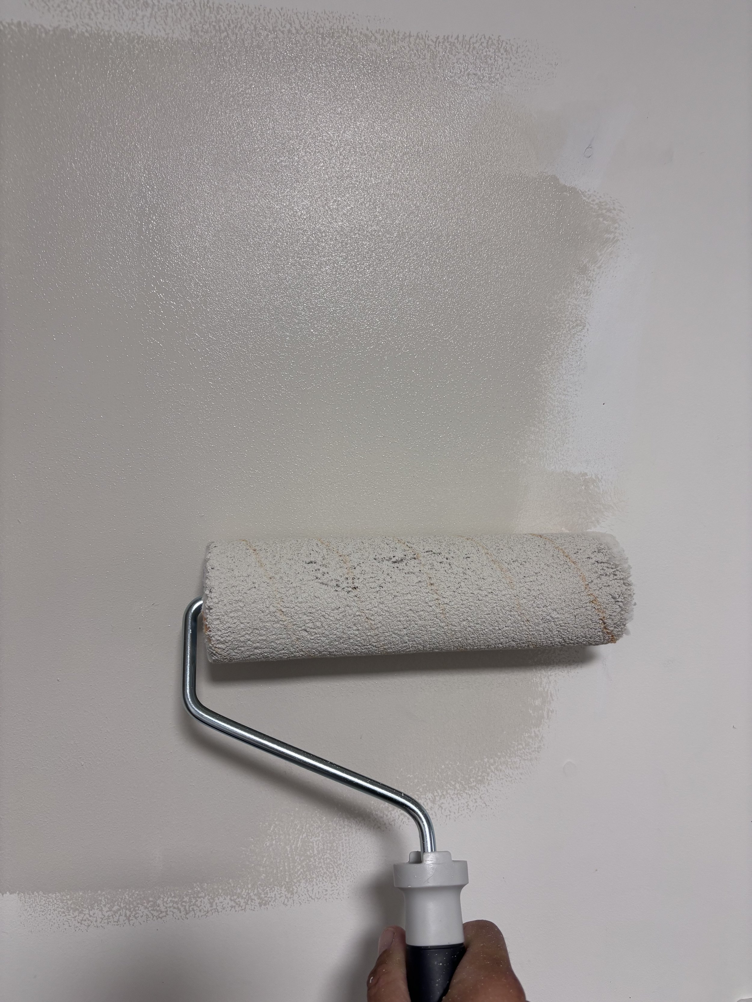 A hand holding a paint roller with a white handle, applying white paint to a wall.