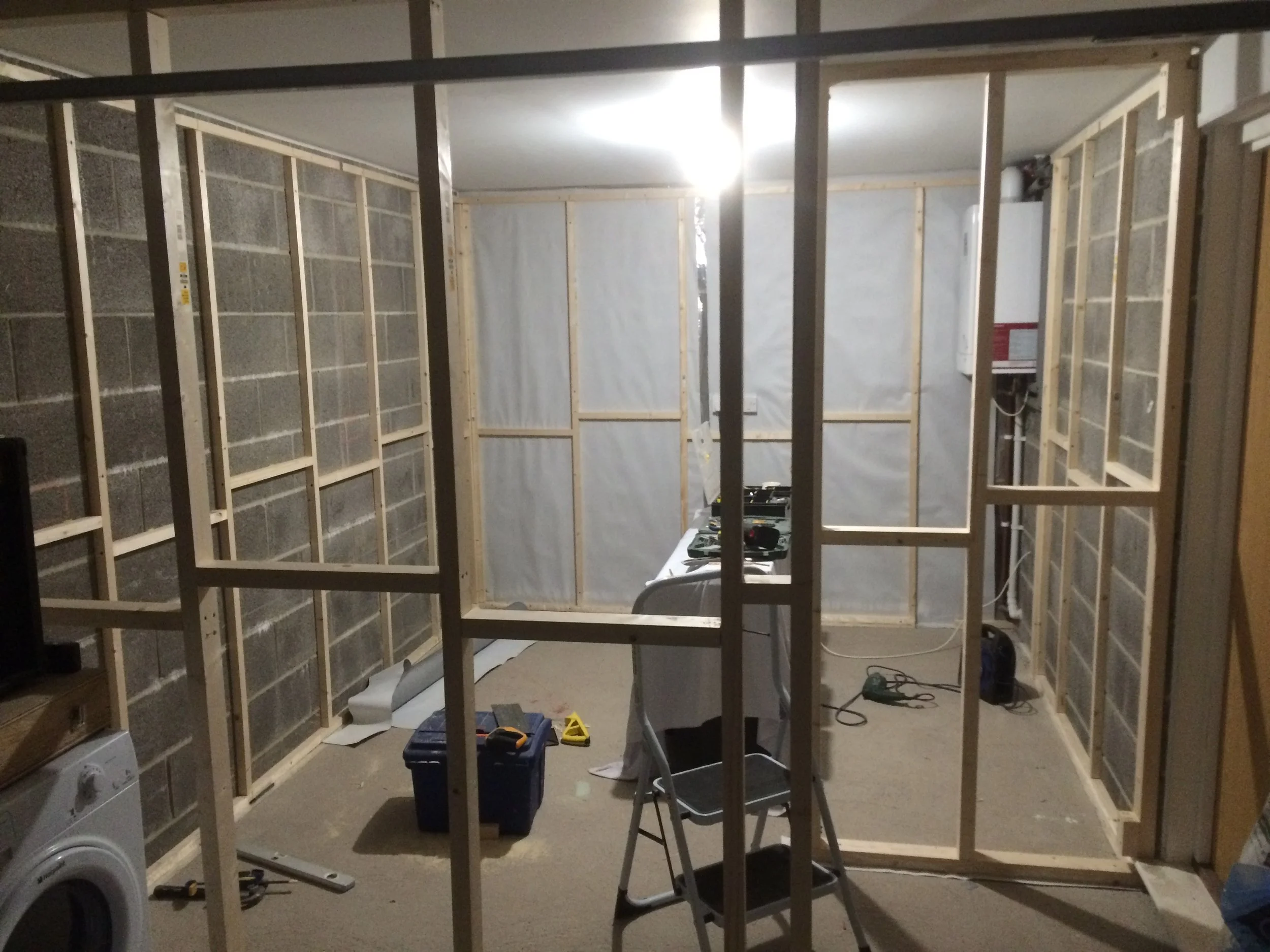 Construction site with unfinished wooden wall framing in progress, tools and supplies on the floor, and electrical outlets installed, with a bright overhead light