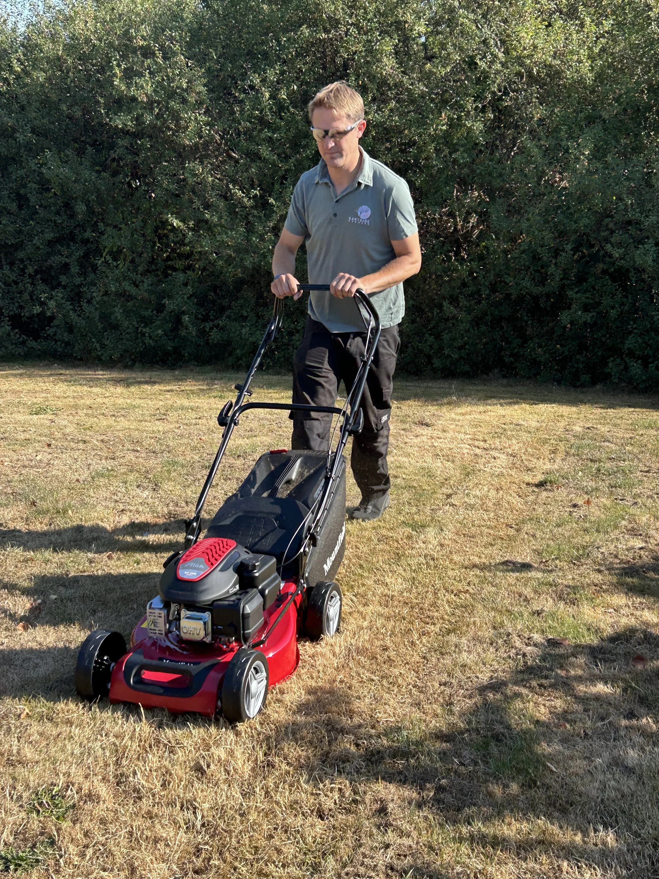A person in sunglasses pushing a red and black lawn mower on a grassy lawn with trees in the background.