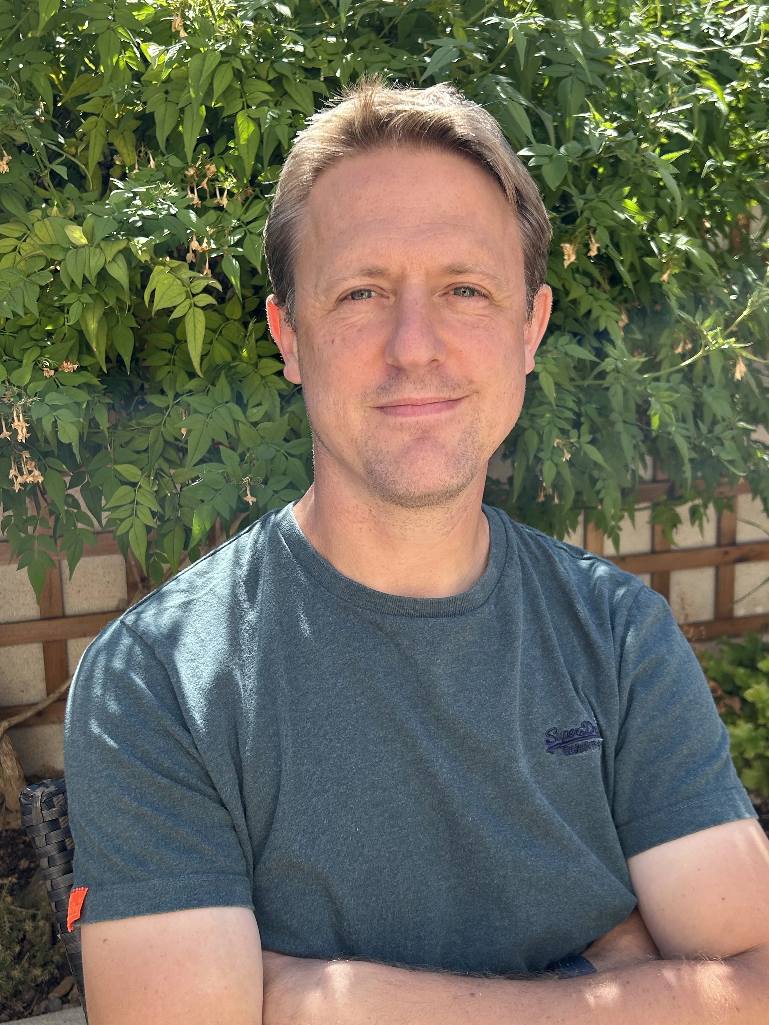 A man with short brown hair and blue eyes smiling, sitting outdoors in front of green leafy bushes with small beige flowers.