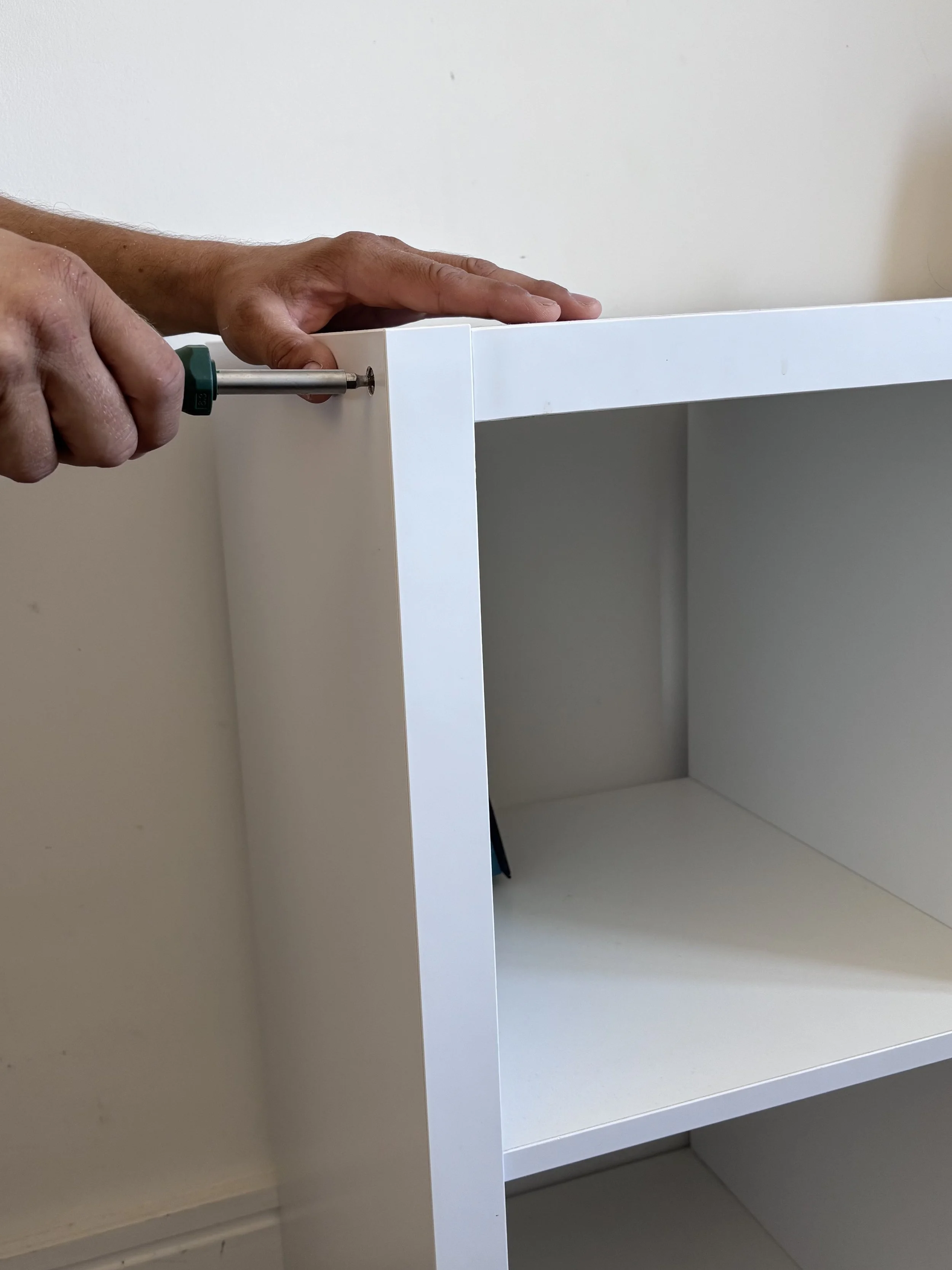 A person using a screwdriver to assemble or tighten a corner of a white wooden cabinet or shelf unit.