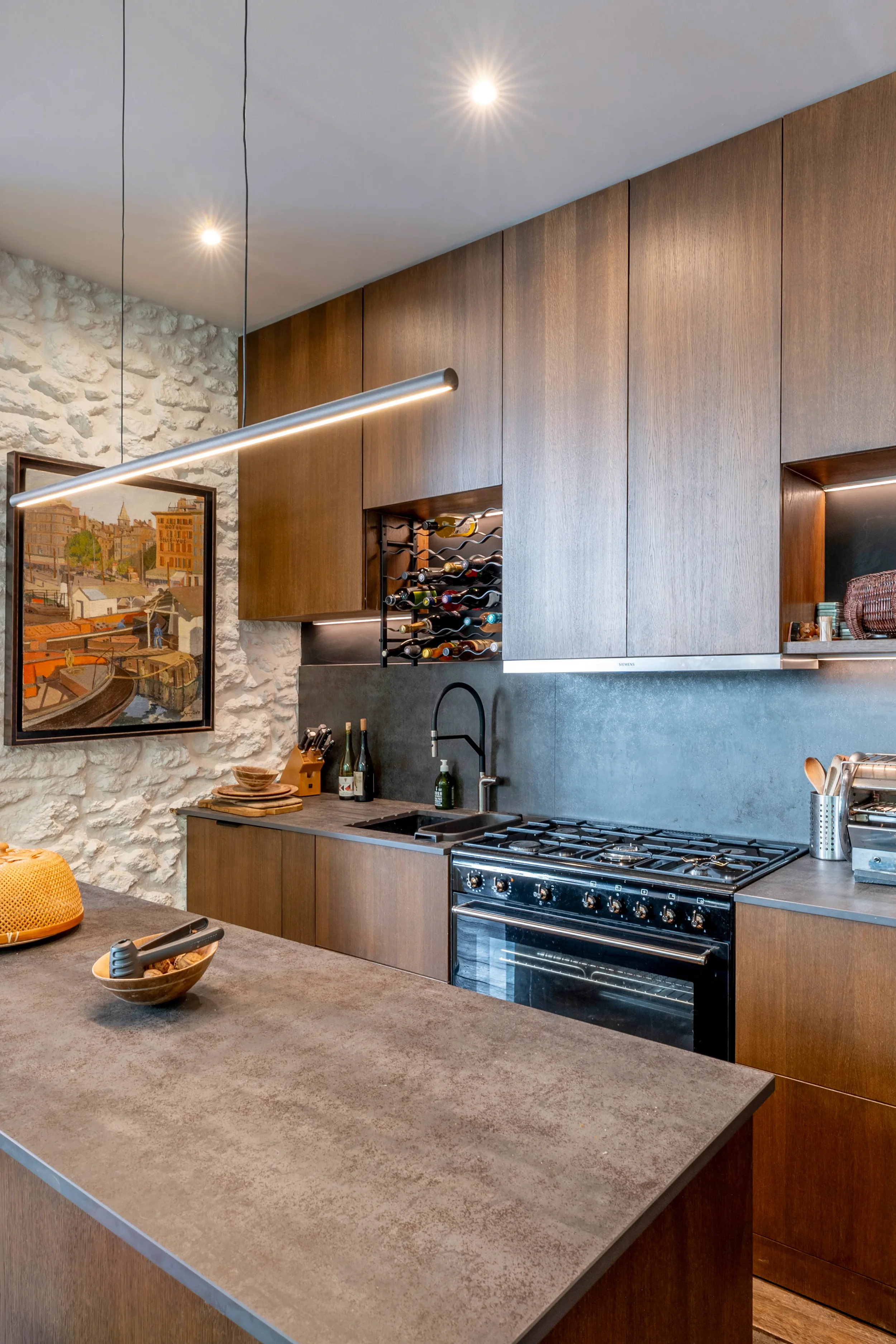 Cuisine haut de gamme sur-mesure en bois sombre avec plan de travail minéral, mur en pierres apparentes rétroéclairé et luminaire linéaire suspendu