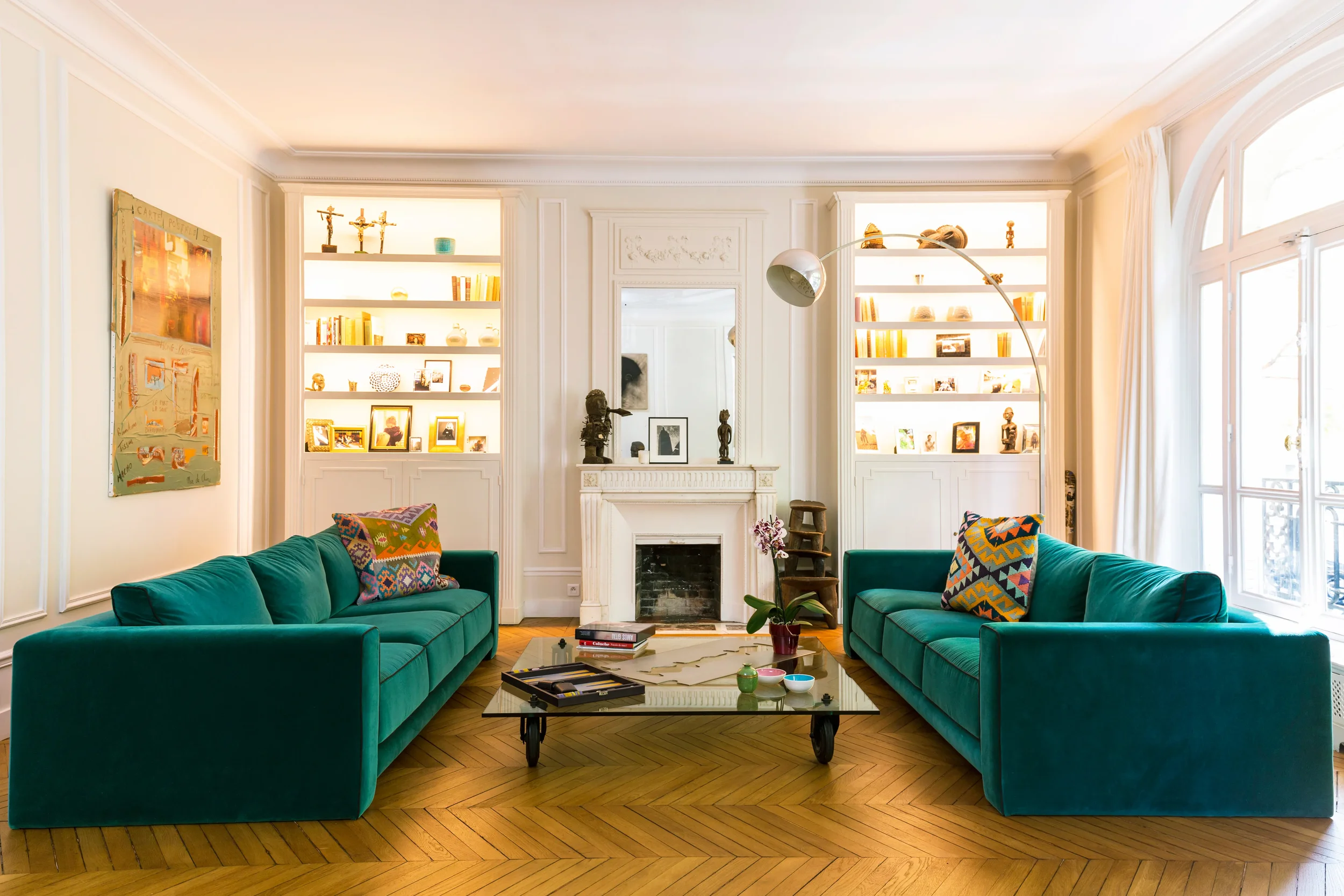 Salon avec deux canapés verts, une cheminée blanche, étagères avec des objets décoratifs et des photos, grande fenêtre laissant entrer la lumière, tapis, table basse avec quelques objets.