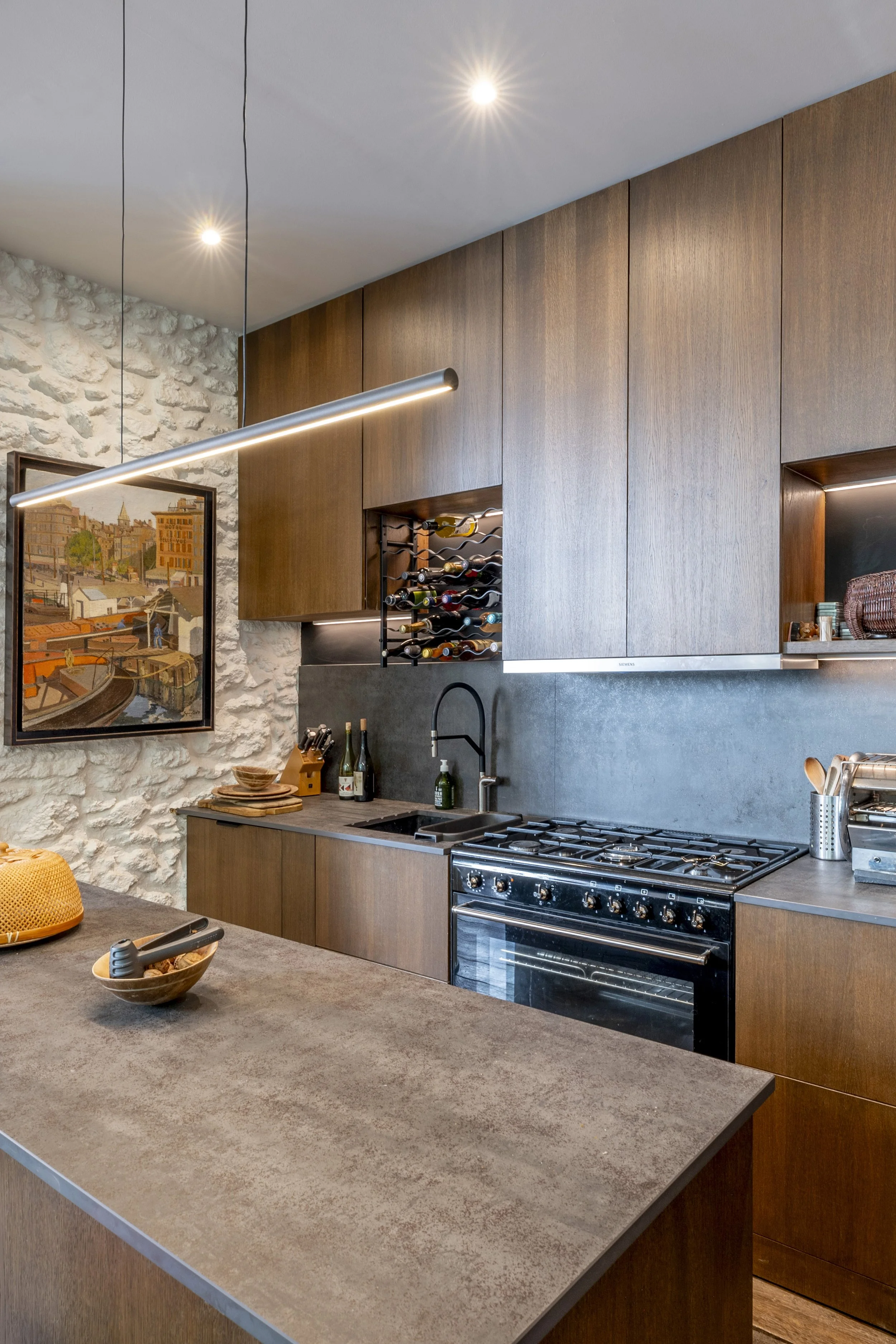Cuisine sur-mesure en bois foncé avec îlot en pierre, mur en moellons et îlot central à Lamarck Caulaincourt par DMM Interiors.