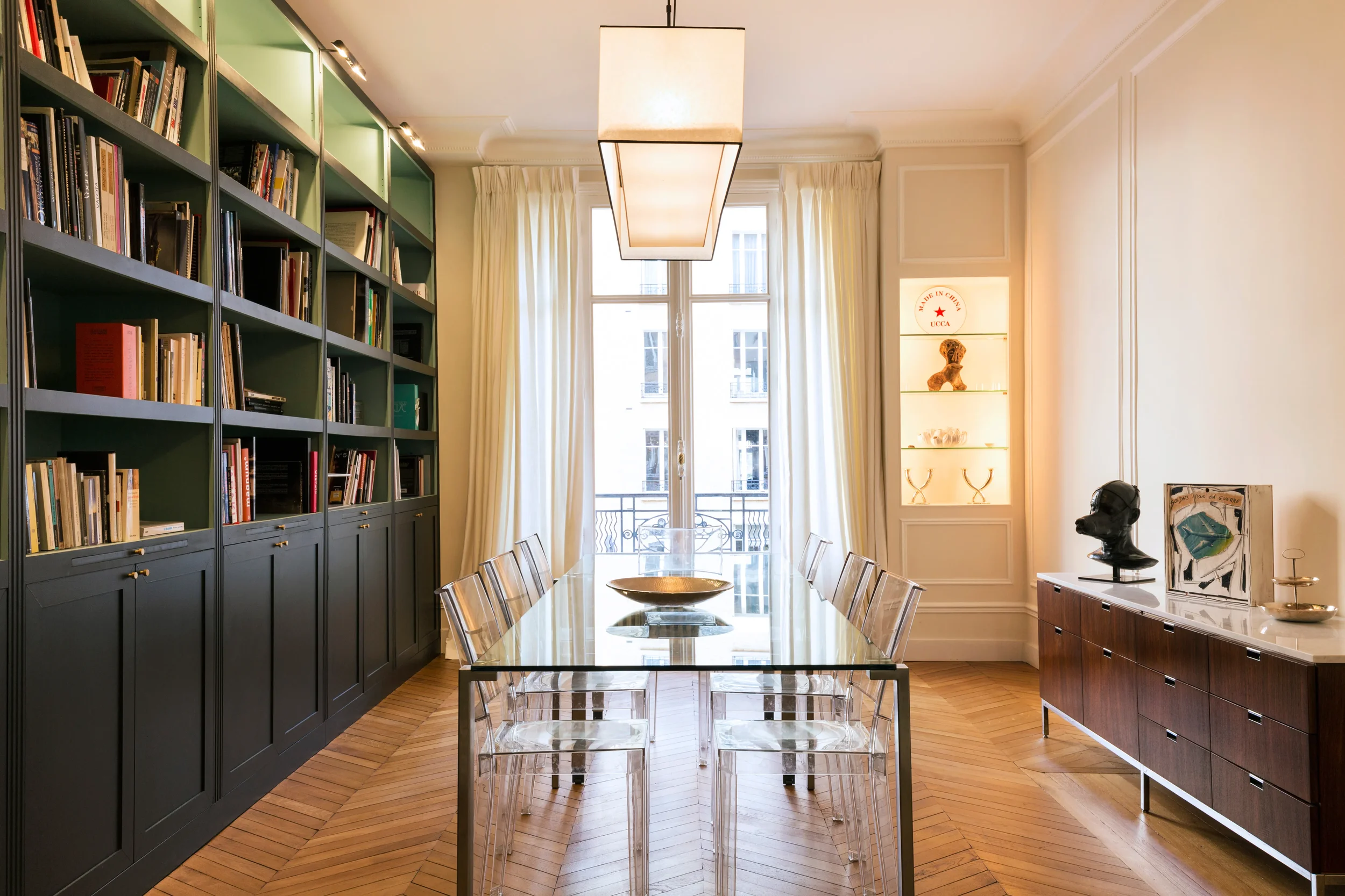 Salle à manger avec bibliothèque sur-mesure sombre, table en verre, chaises Ghost et parquet Point de Hongrie, projet Neuilly.