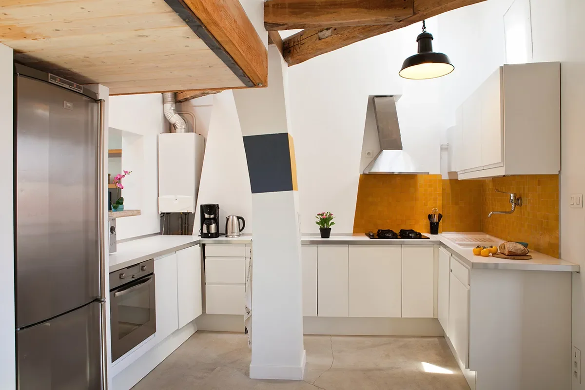 Cuisine ouverte sur-mesure aux meubles blancs, crédence en zelliges jaune safran, poutres en bois brut au plafond et poteau central dans un loft à Bastille